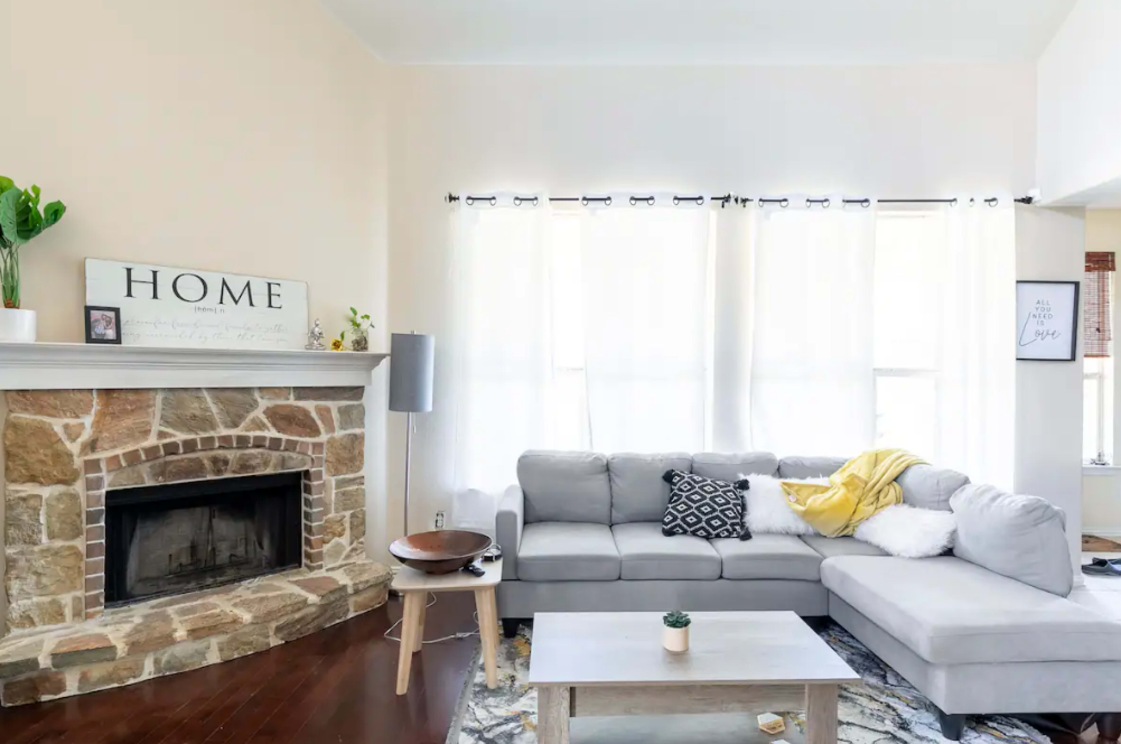 The living room features a stone fireplace, a gray sectional sofa, and a light-colored coffee table, with large windows allowing natural light to fill the space.