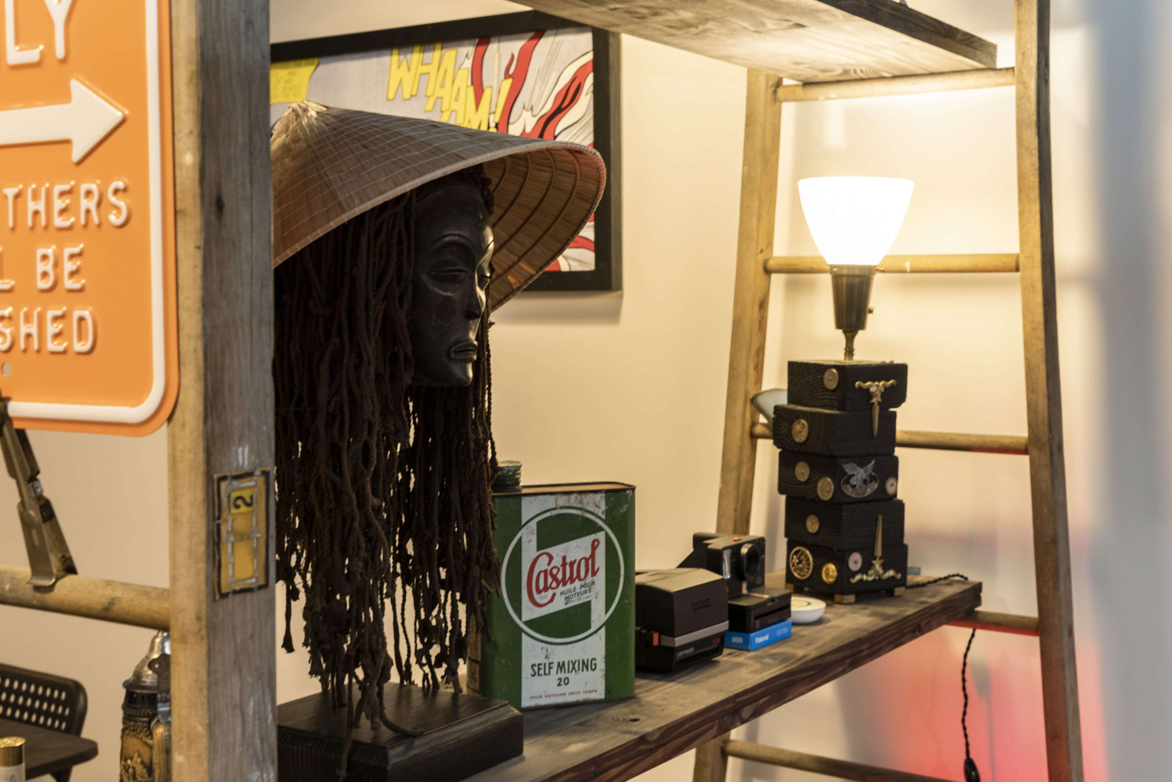 A wooden shelf displays a decorative bust with a hat, a vintage can, and various small boxes under a lamp.