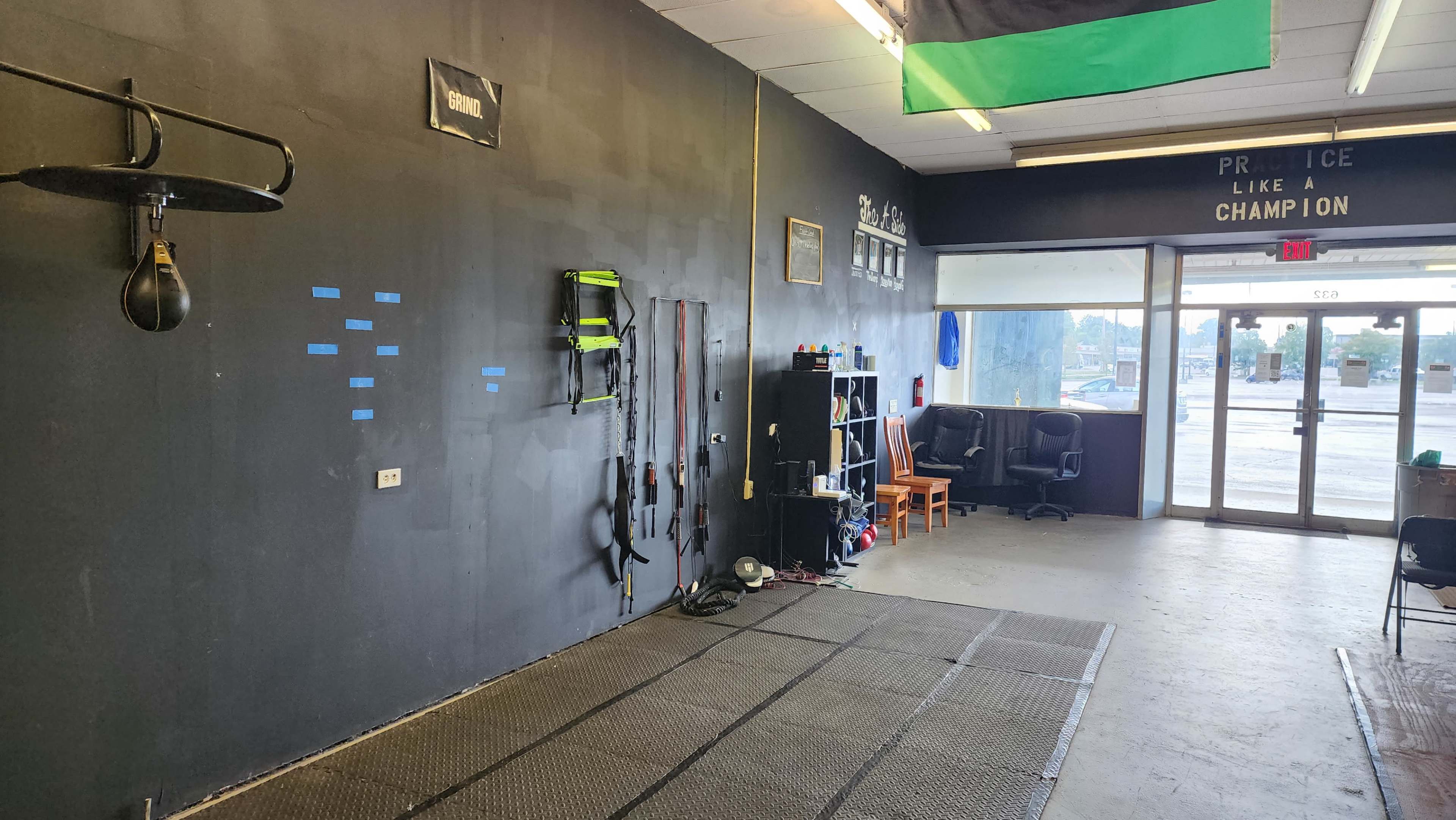 A sparsely furnished training space featuring a boxing speed bag hanging on the left wall, various workout equipment organized on a black shelf, and a large window at the entrance revealing outdoor light.