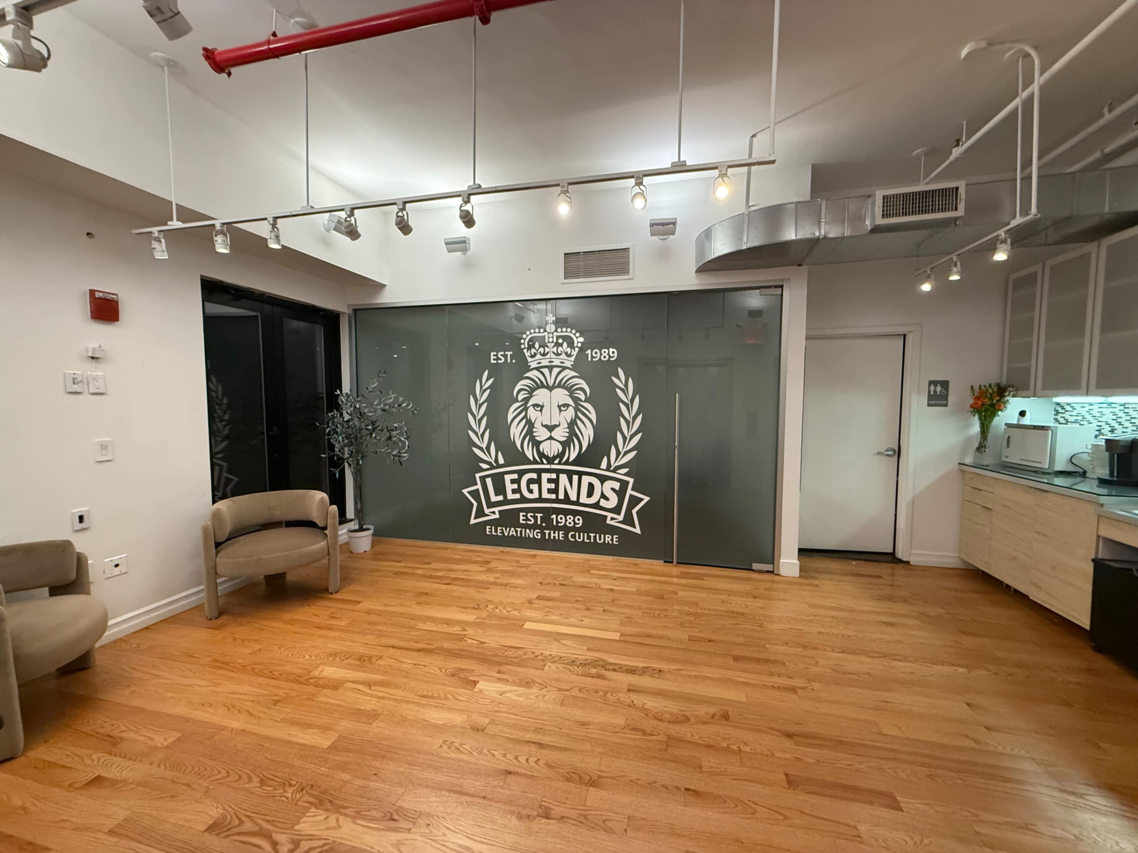 The image shows a modern interior space with wooden flooring, featuring a glass wall adorned with a lion logo and the text "LEGENDS EST. 1989," along with two tan chairs.
