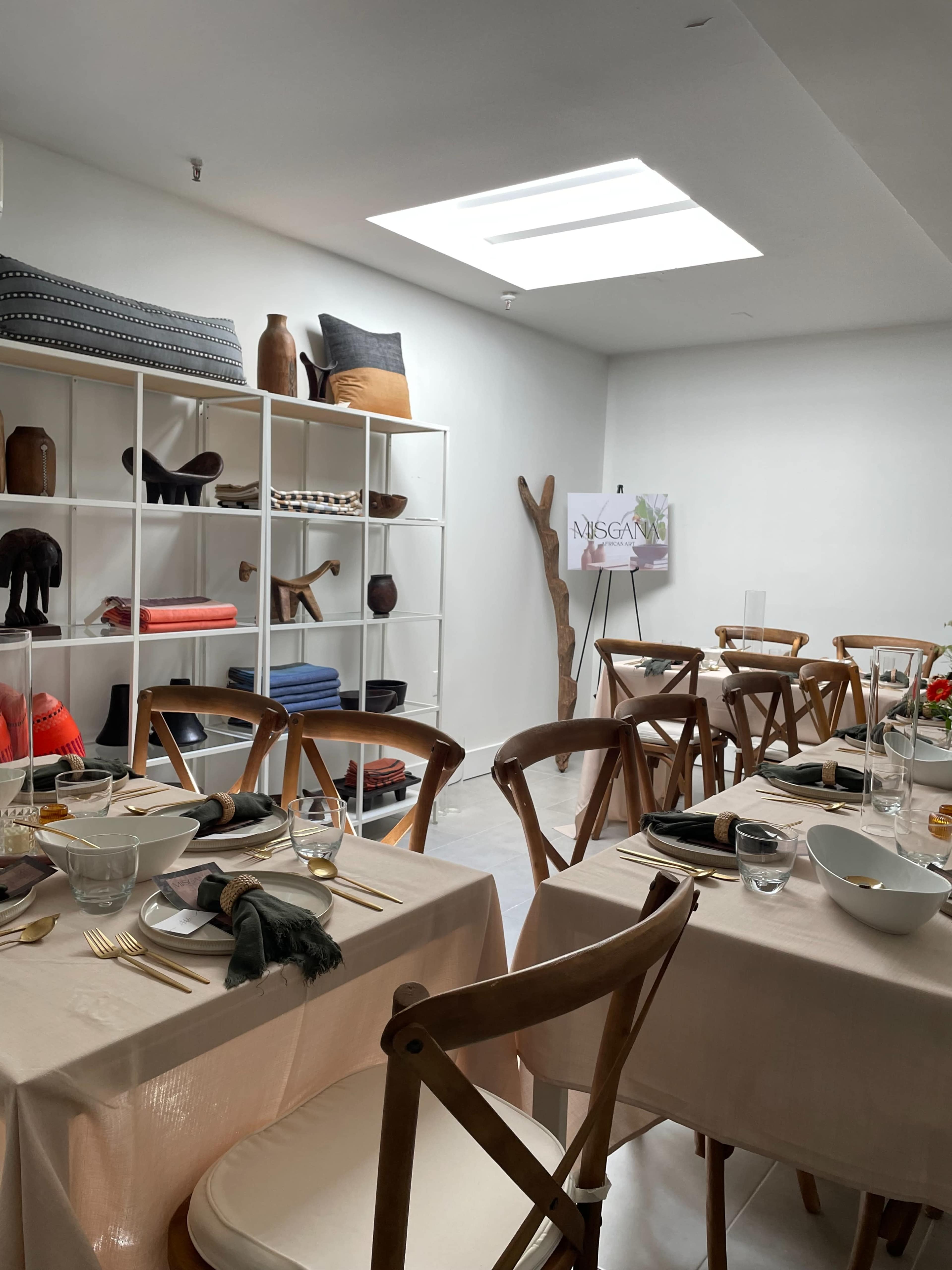 The image depicts a minimally decorated dining area with wooden tables set for a meal, surrounded by shelves displaying various decorative items.