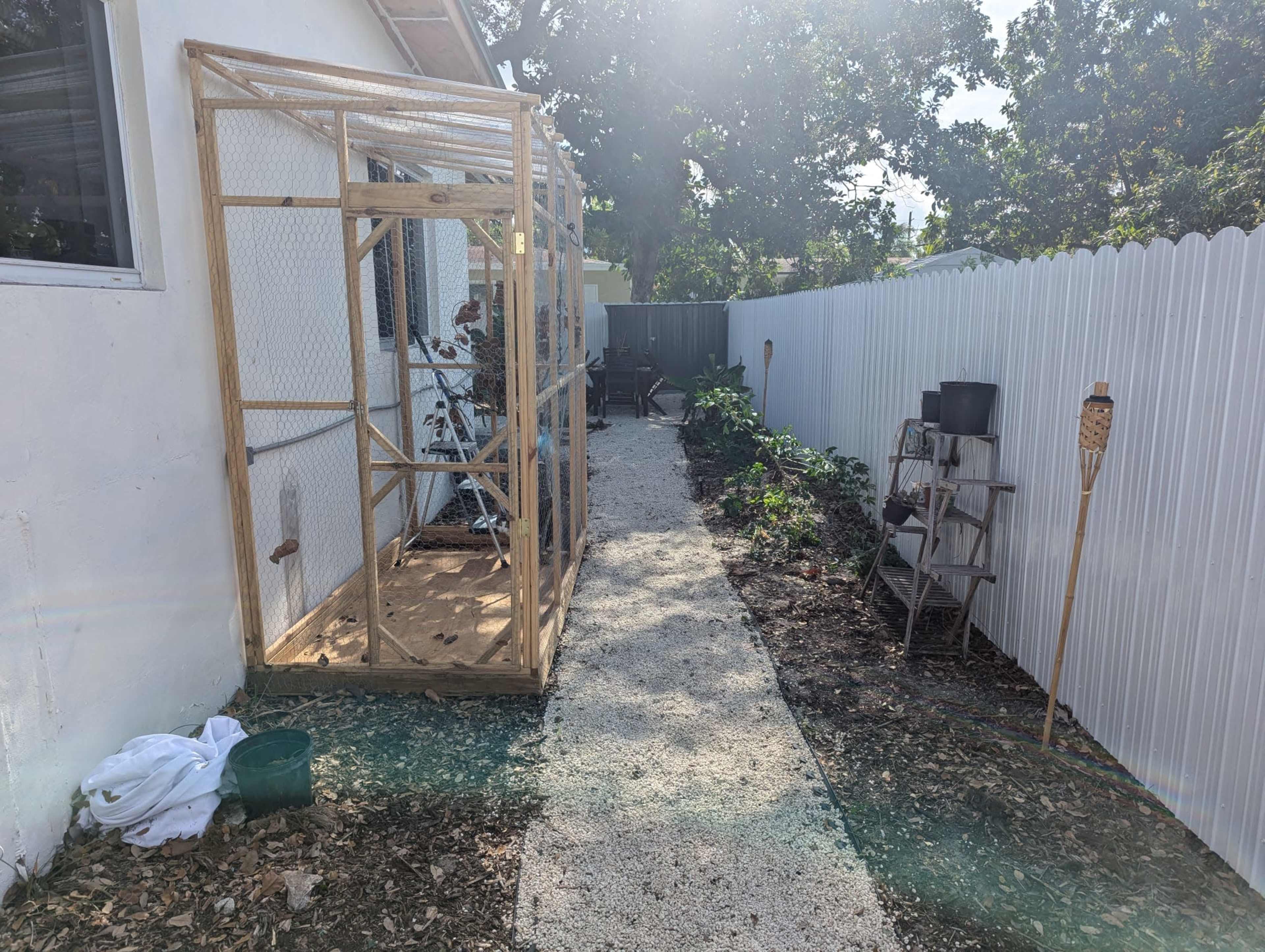 A wooden frame structure is positioned along a gravel path beside a white fence, with plants and a small garden area visible on one side.