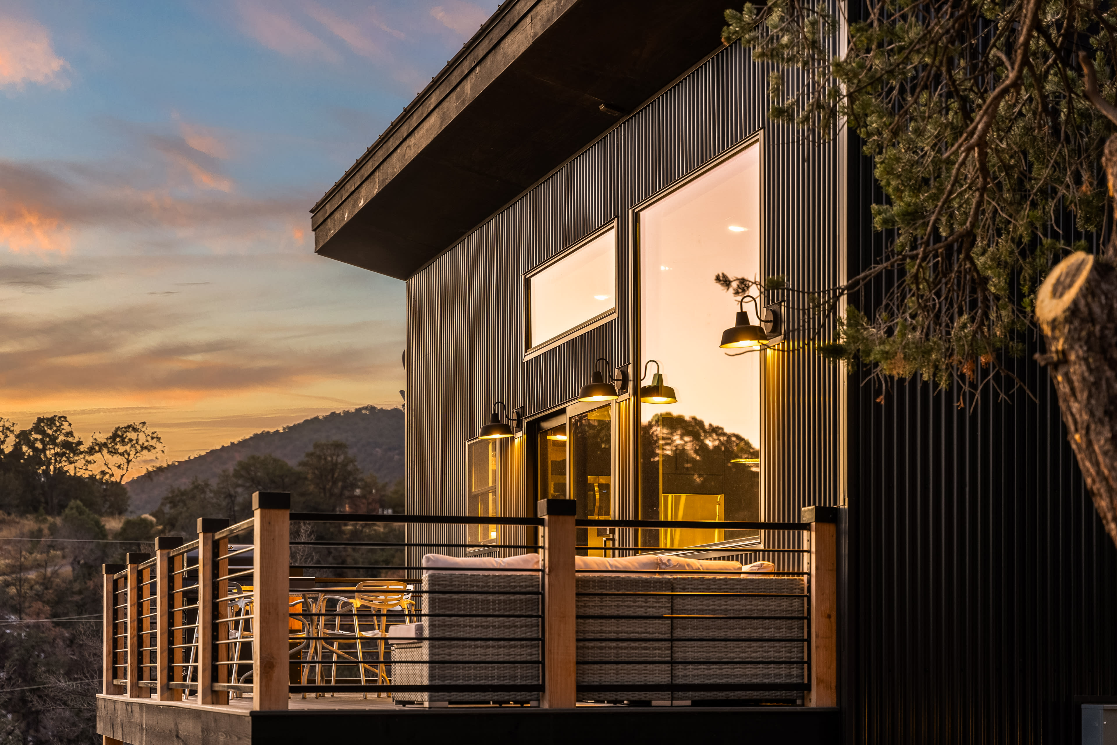 A modern cabin with large windows and a balcony overlooks a scenic sunset landscape.