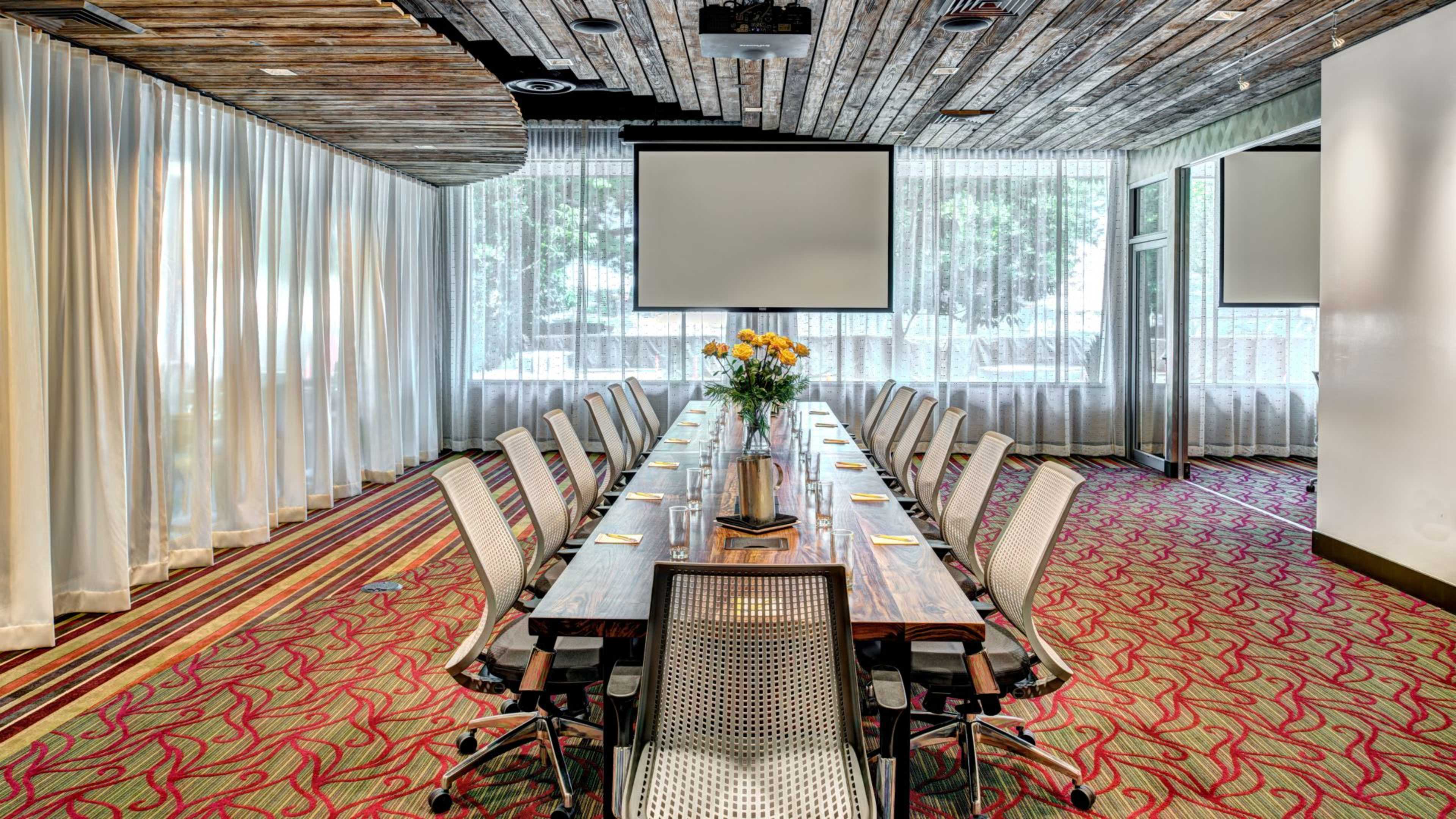 A long wooden conference table is set with water and notebooks, surrounded by ergonomic chairs, in a bright meeting room with large windows and sheer curtains.