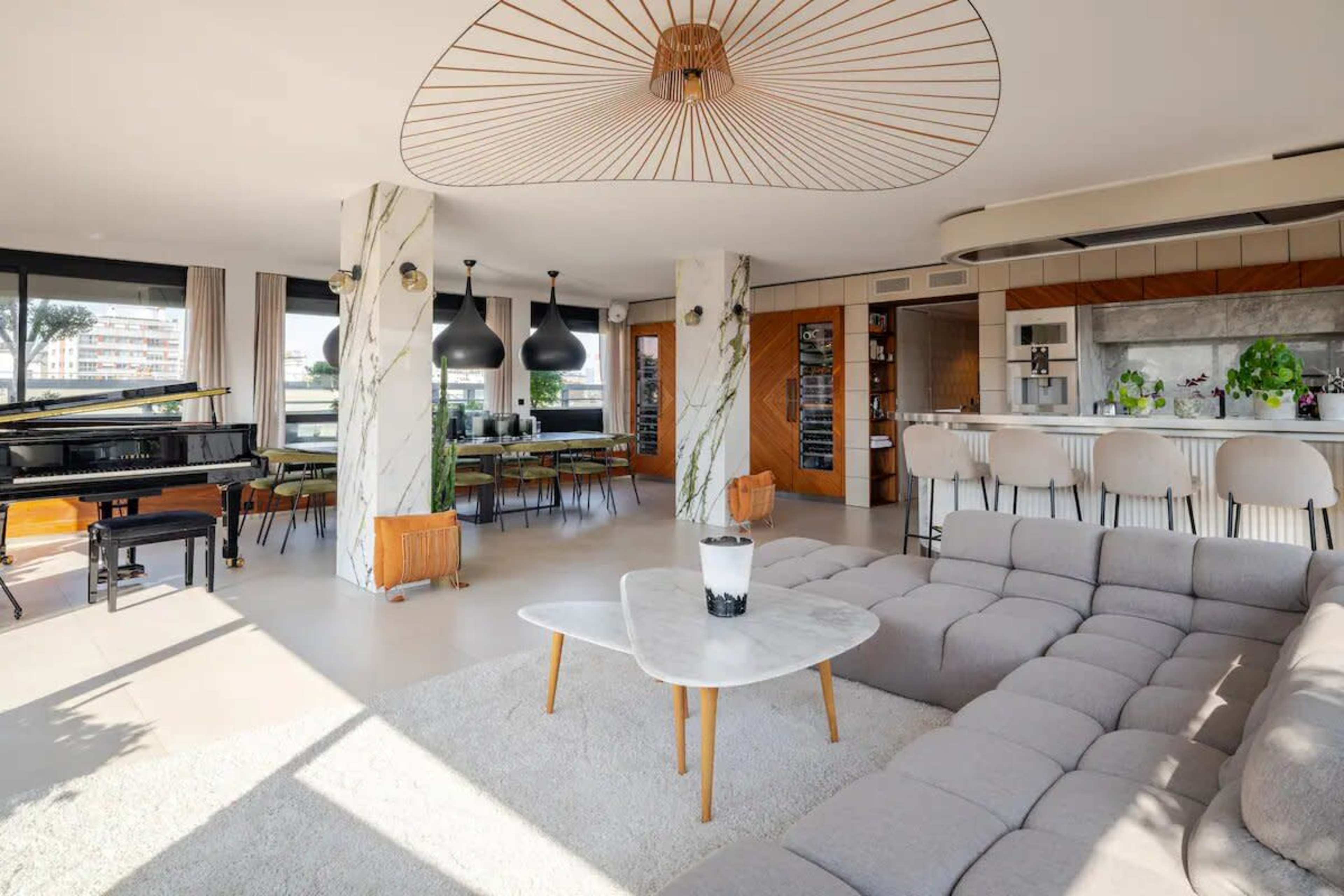A modern open-concept living space featuring a gray sectional sofa, a marble coffee table, a grand piano, and a kitchen with bar seating.