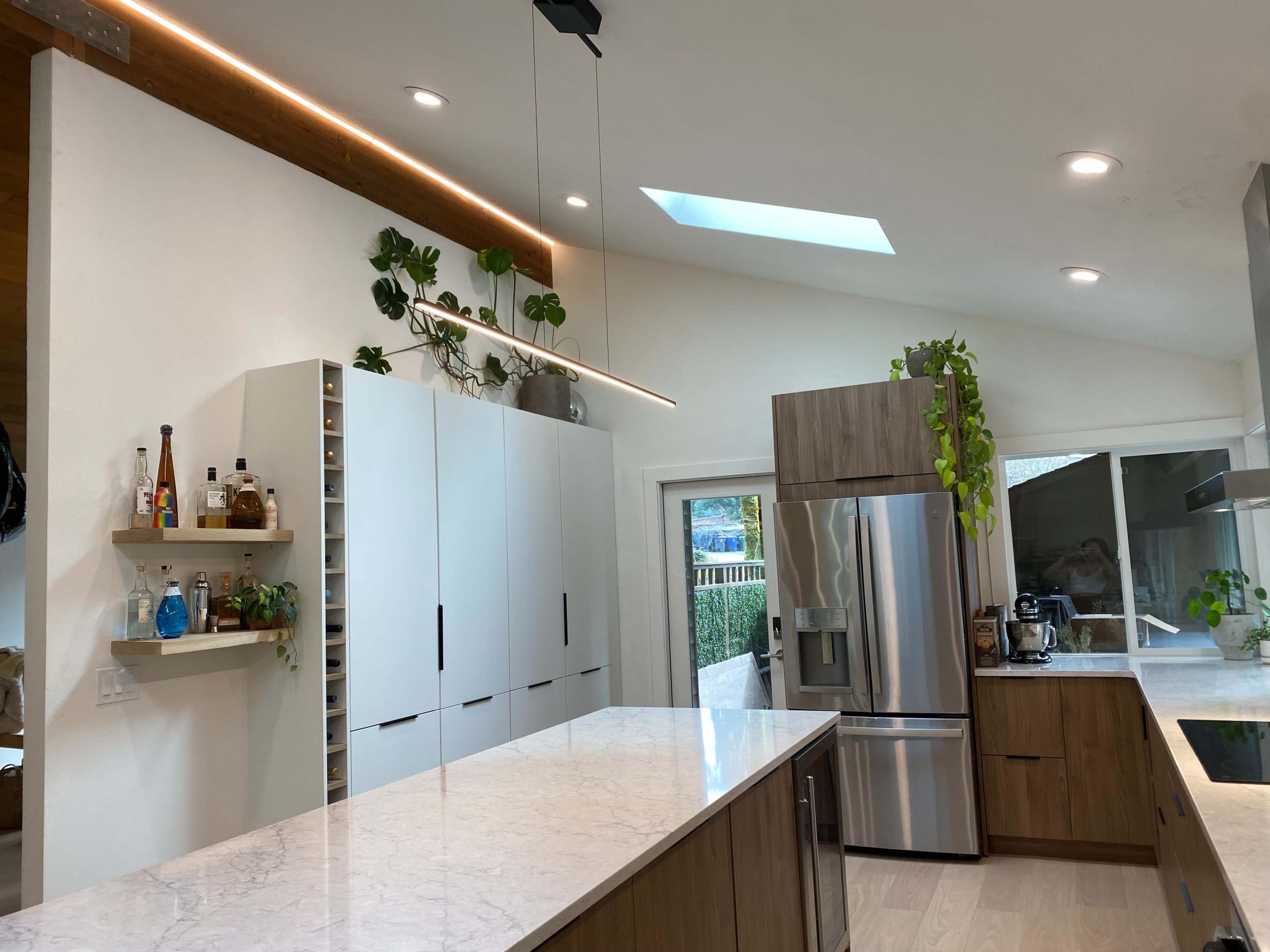 A modern kitchen with light wood cabinetry, a large marble island, and plants placed along the shelves and walls.