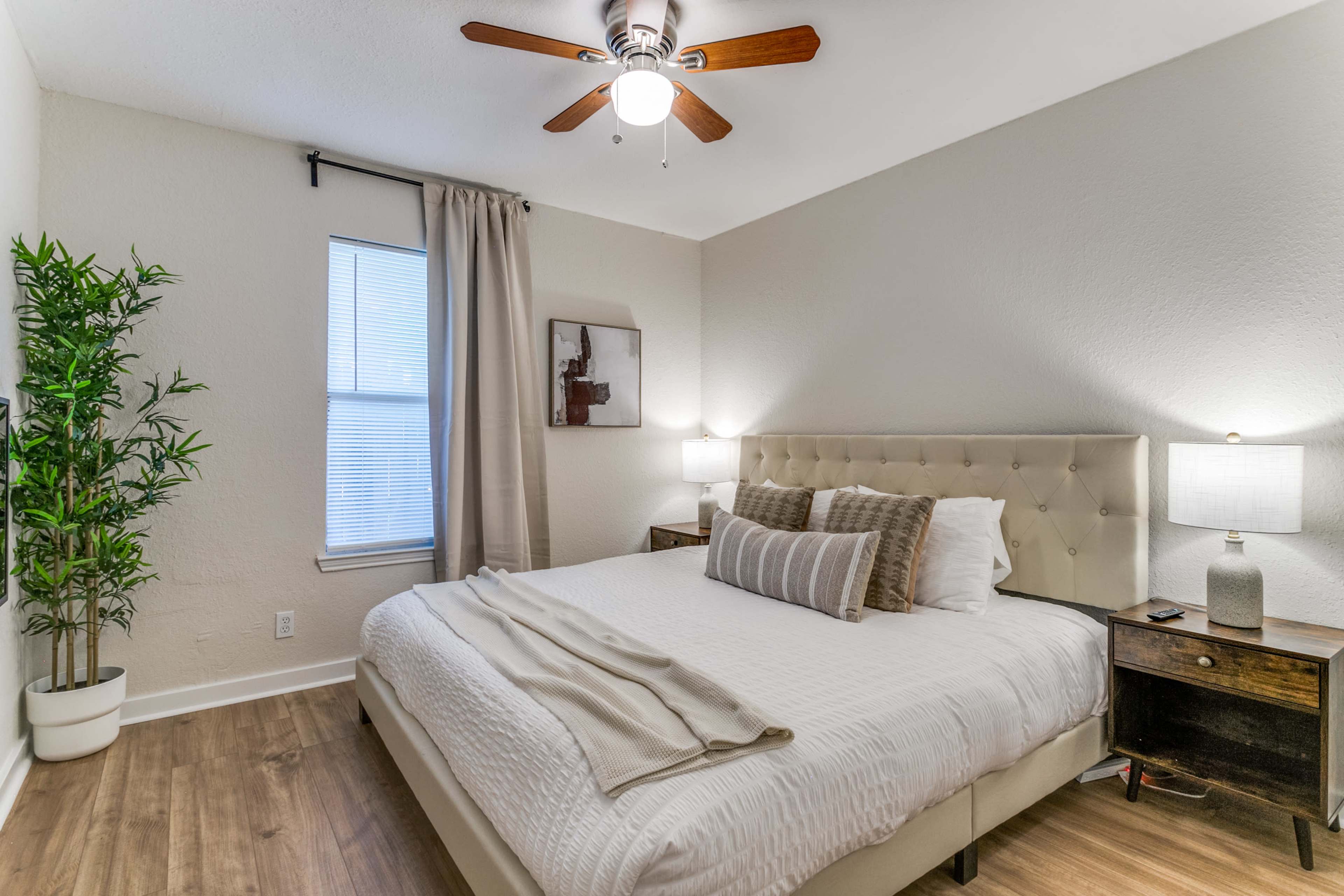 A neatly arranged bedroom features a large bed with neutral bedding, side tables with lamps, and a window beside a potted plant.