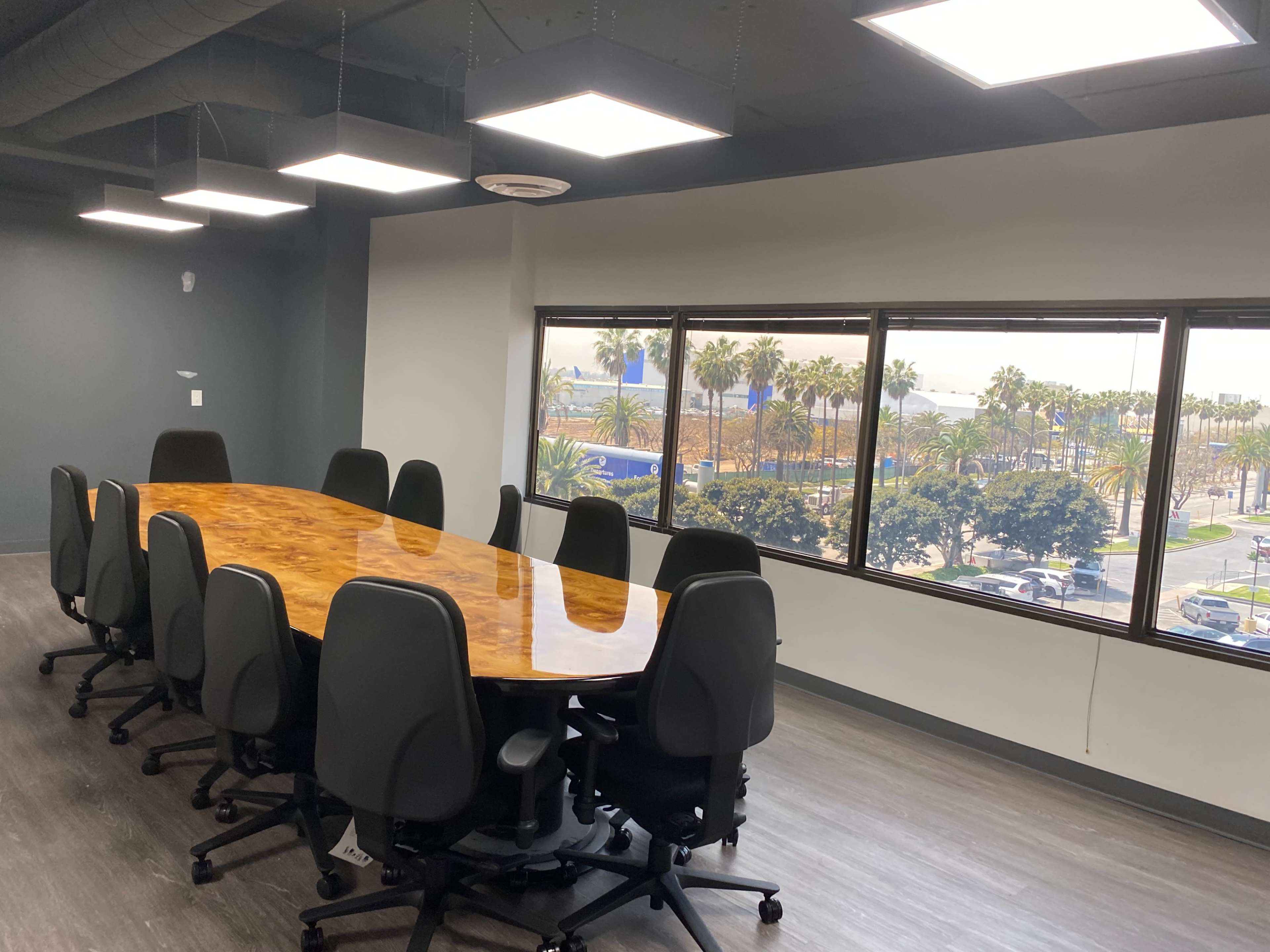 A long wooden conference table surrounded by executive chairs sits in a bright meeting room with large windows overlooking palm trees and a cityscape.
