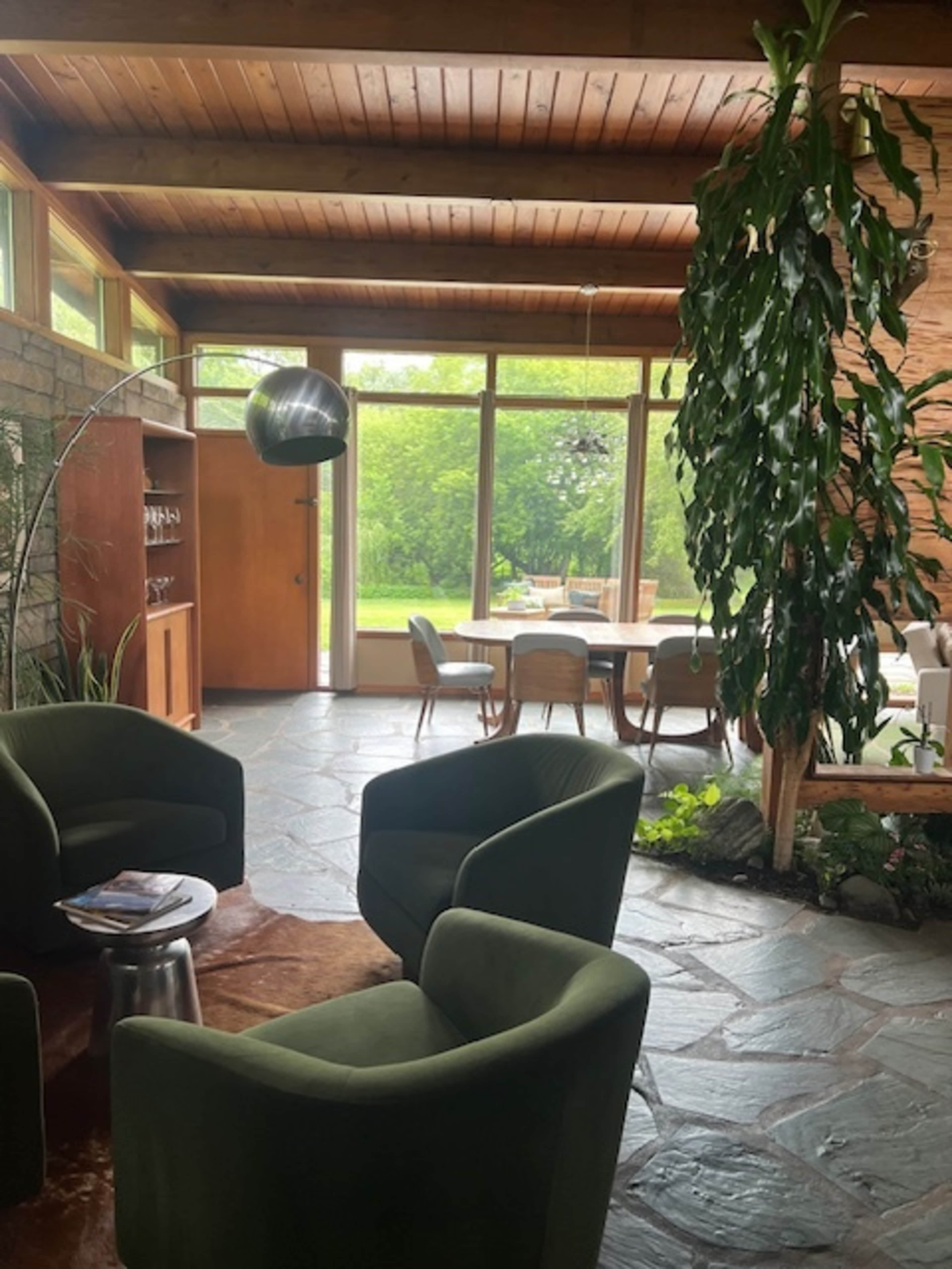 A spacious living area features two green armchairs, a stone floor, and large windows overlooking a garden.