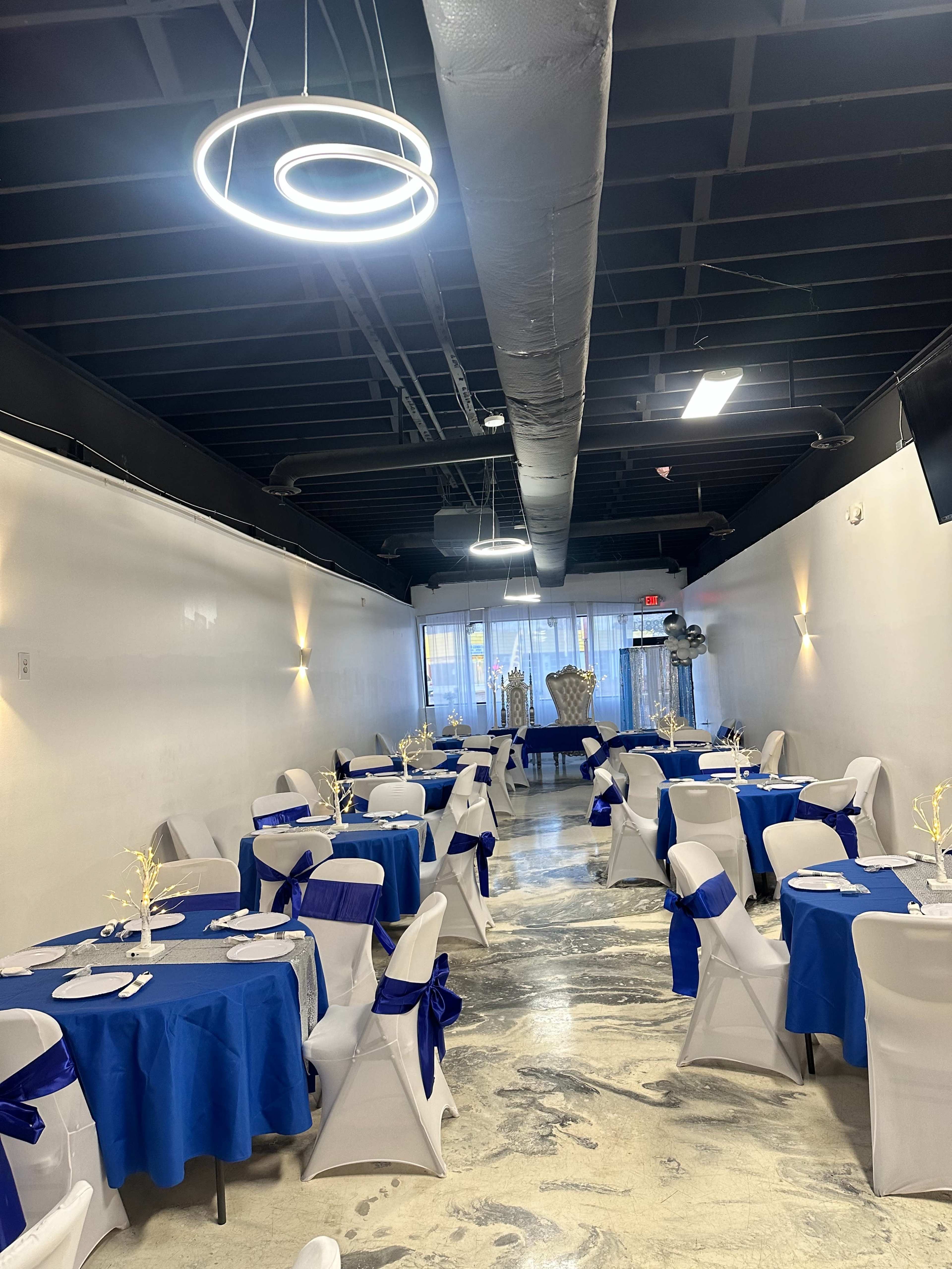 The image shows a spacious dining area with white walls, blue tablecloths, and elegant seating arrangements, illuminated by modern light fixtures.
