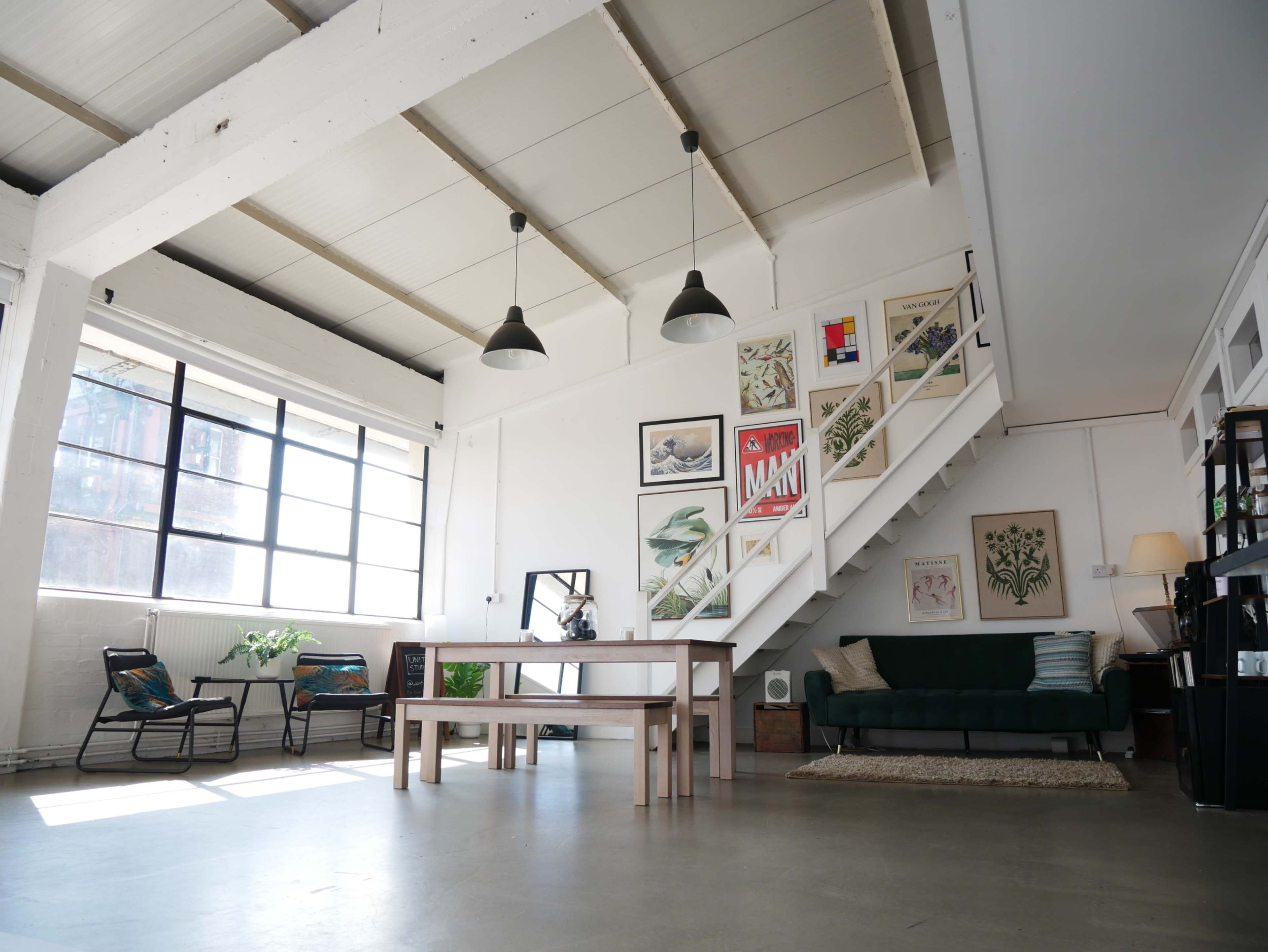 The image shows a spacious, modern interior with a dining table, a green sofa, and art-covered walls, illuminated by large windows.