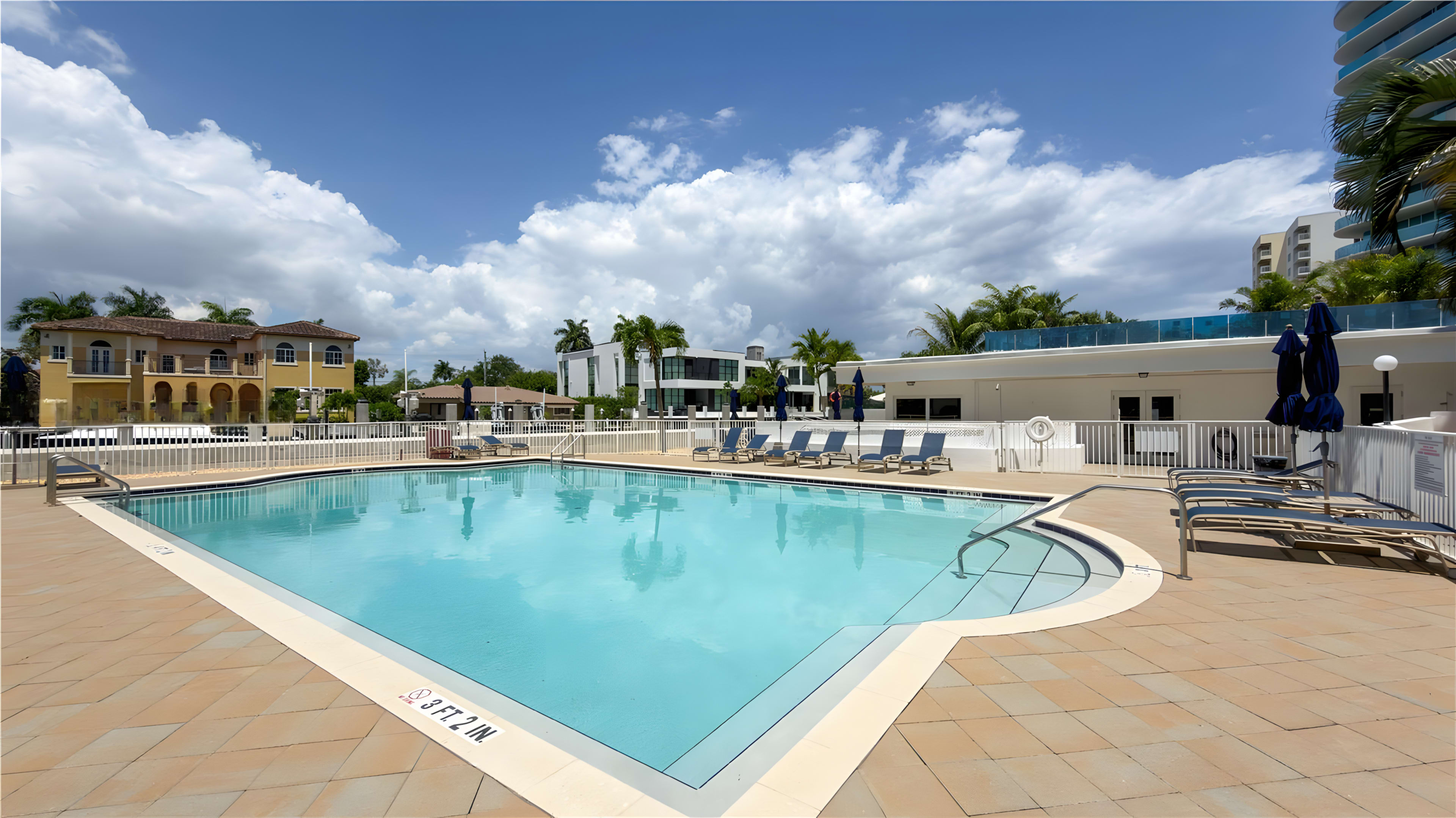 The image shows a clear swimming pool surrounded by lounge chairs and landscaped palm trees, with modern buildings in the background under a partly cloudy sky.
