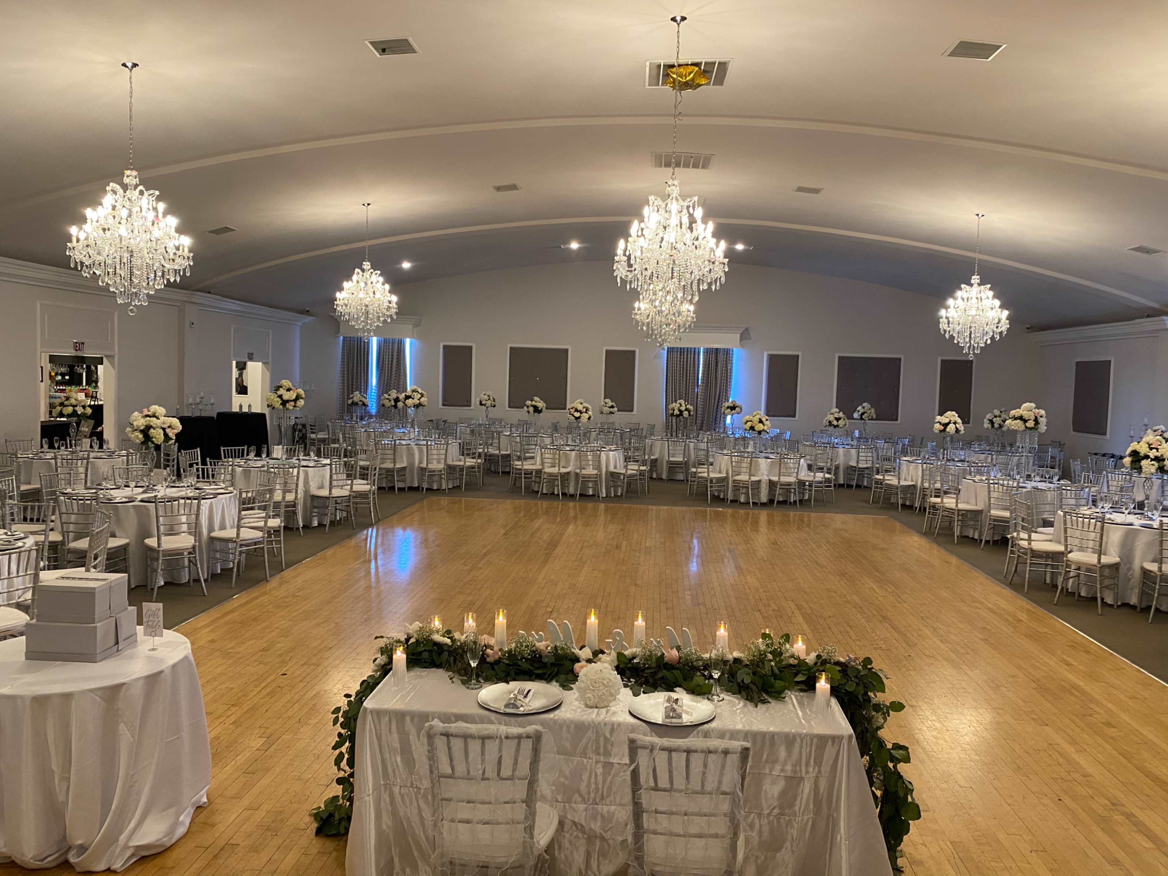 A spacious venue is set up for a formal event, featuring round tables with white linens, elegant chandeliers, and floral centerpieces.