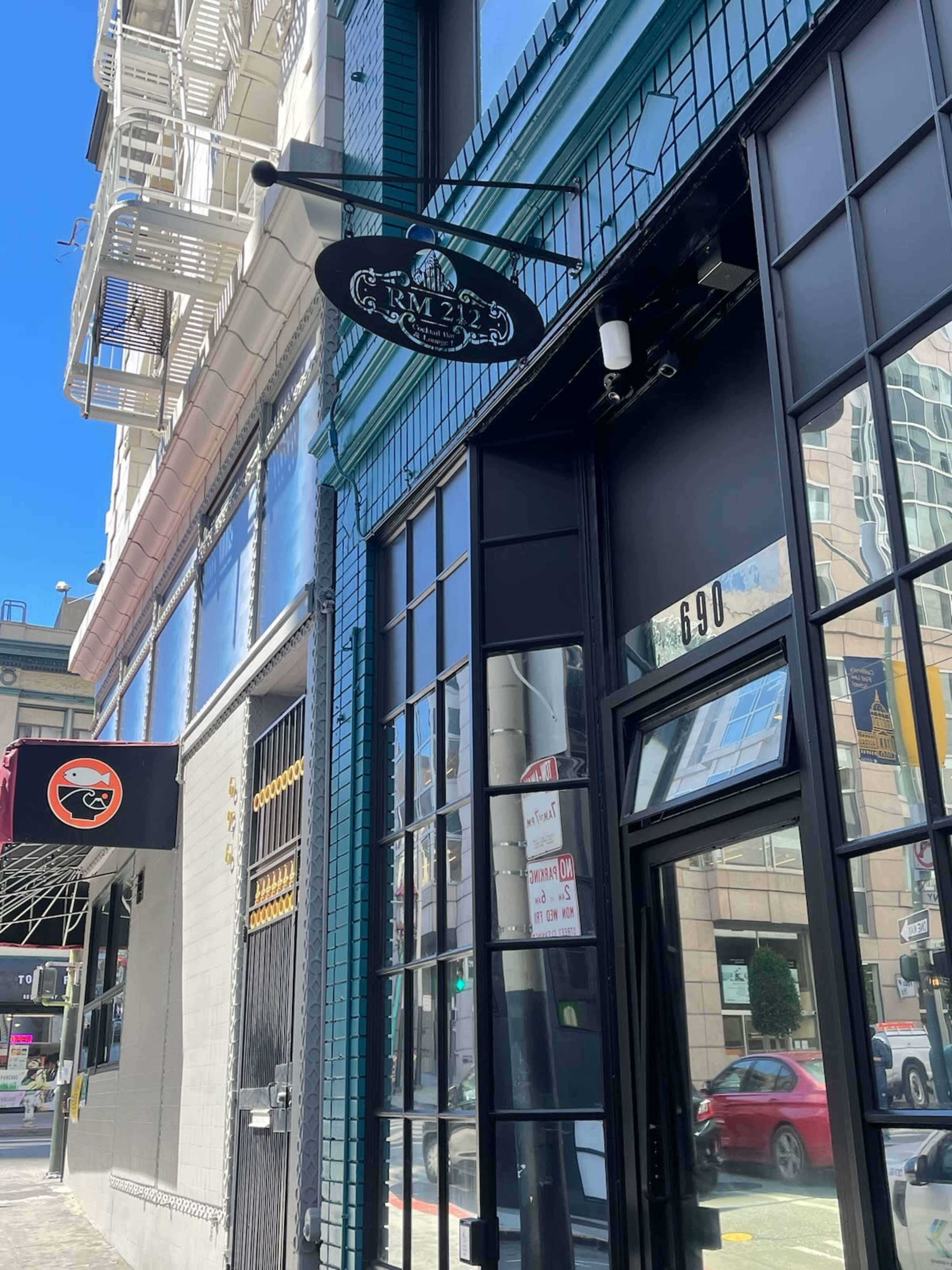 The image shows a bright street scene featuring a storefront with a black sign hanging above the entrance, alongside a building with blue tile accents.