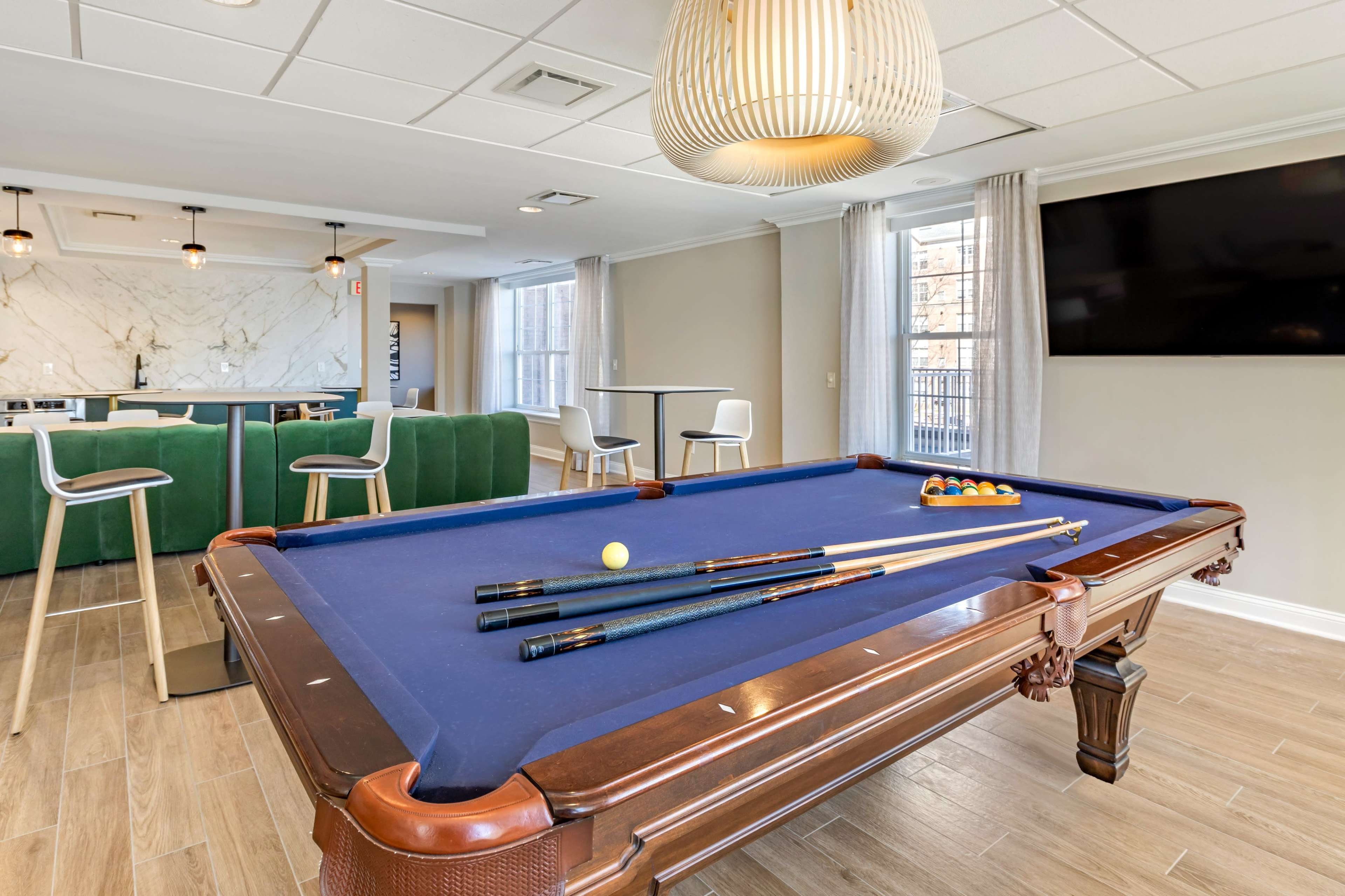 A well-lit game room featuring a billiards table with cues and balls, alongside a seating area and a large television.