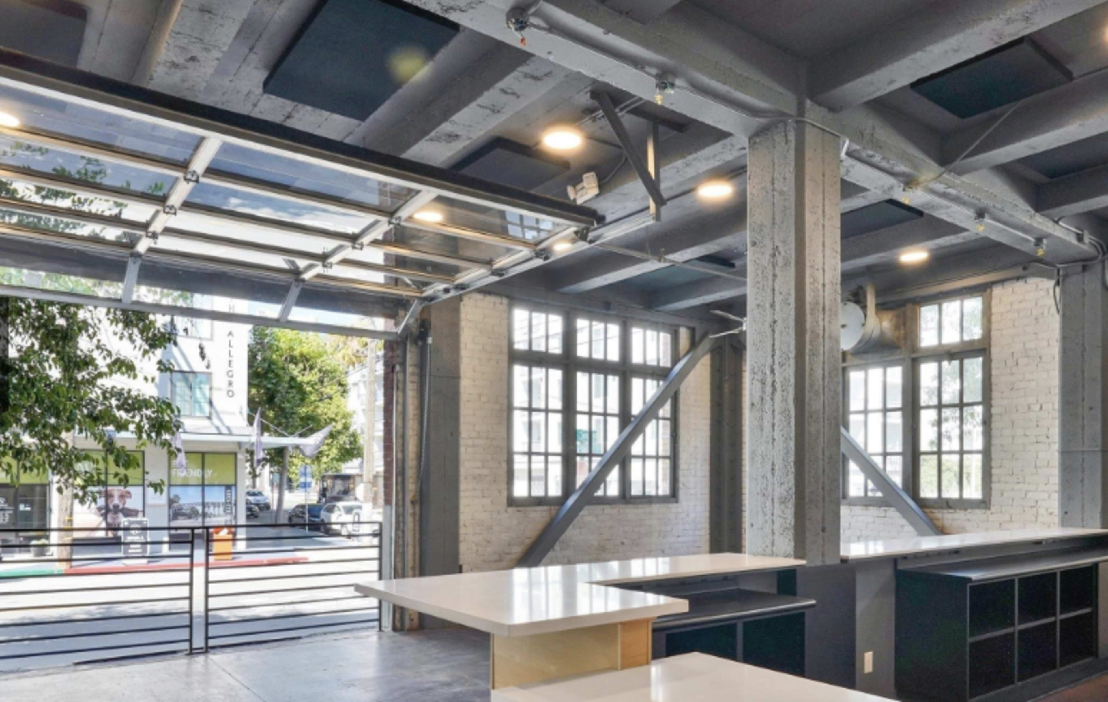 The image shows a modern, open-concept workspace with large windows and a glass garage door, featuring concrete walls and a central island counter.