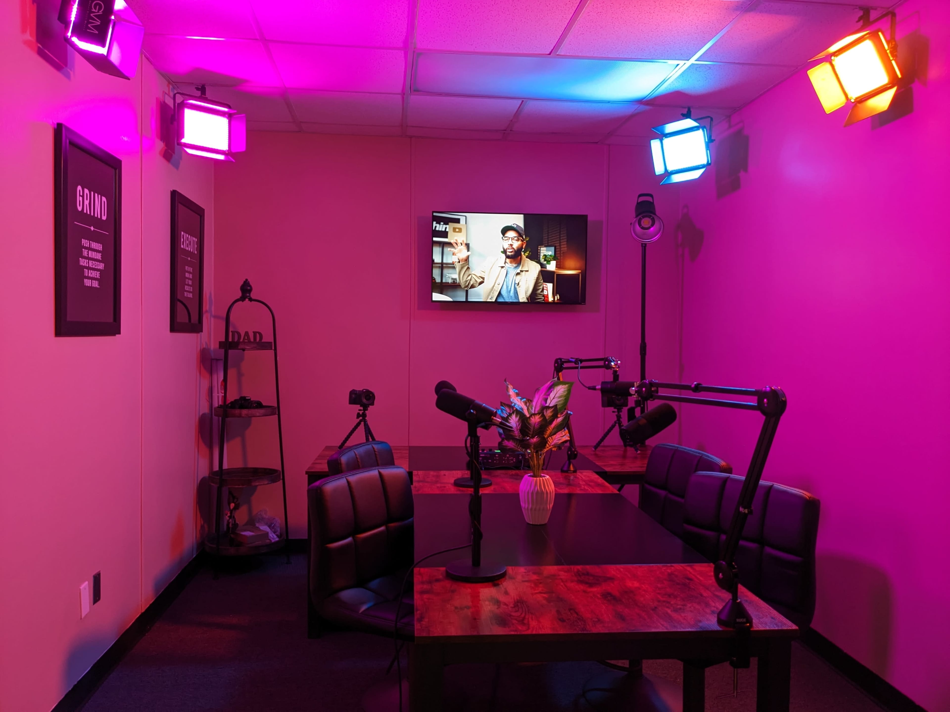 A small recording studio with a table, chairs, and cameras, illuminated by colorful lights and featuring a television screen on the wall.