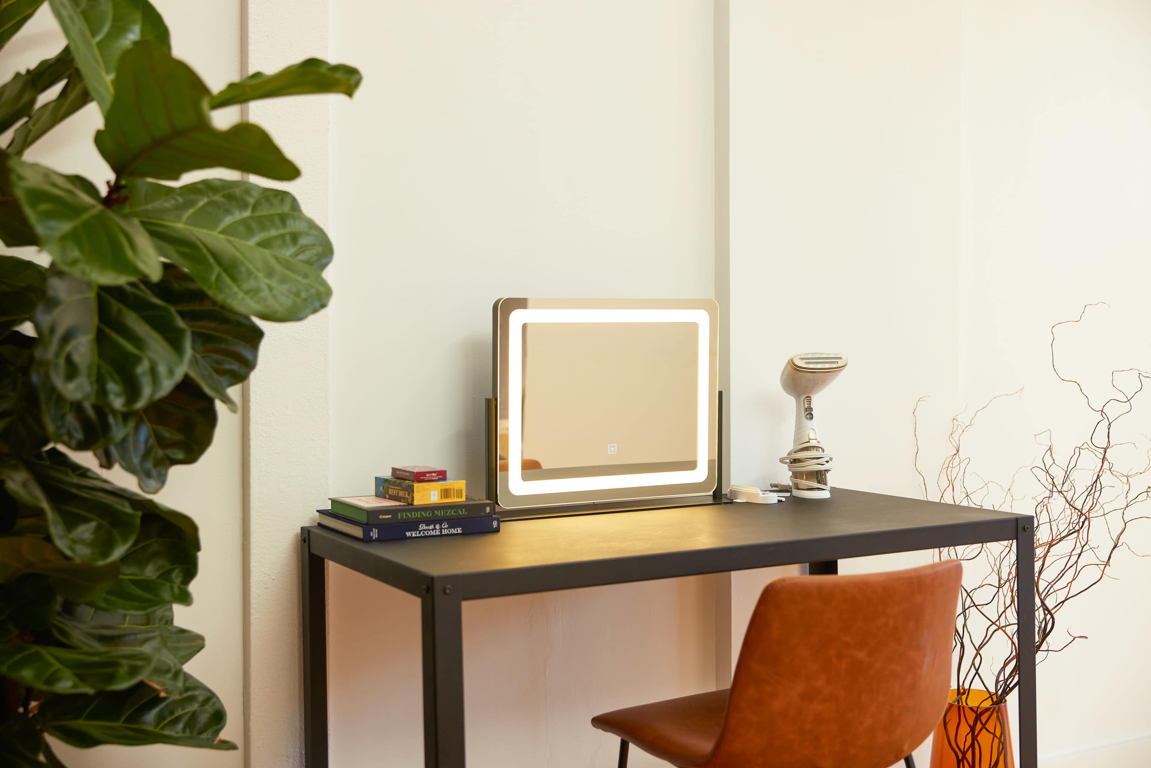 A desk with a lit mirror, surrounded by a large plant and books, features a modern lamp and a decorative vase on the side.