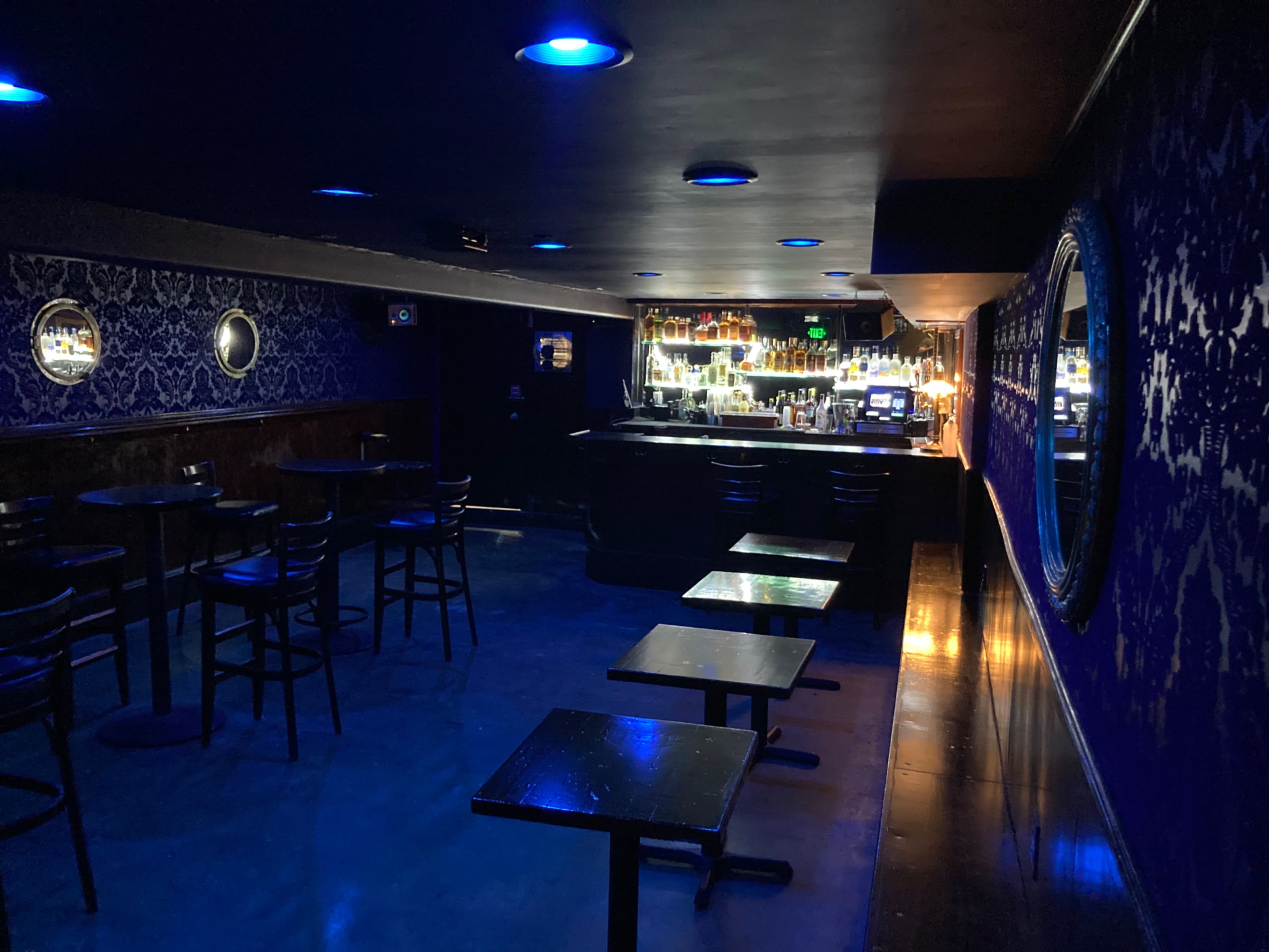 A dimly lit bar with dark walls, scattered tables, and a well-stocked backlit counter.