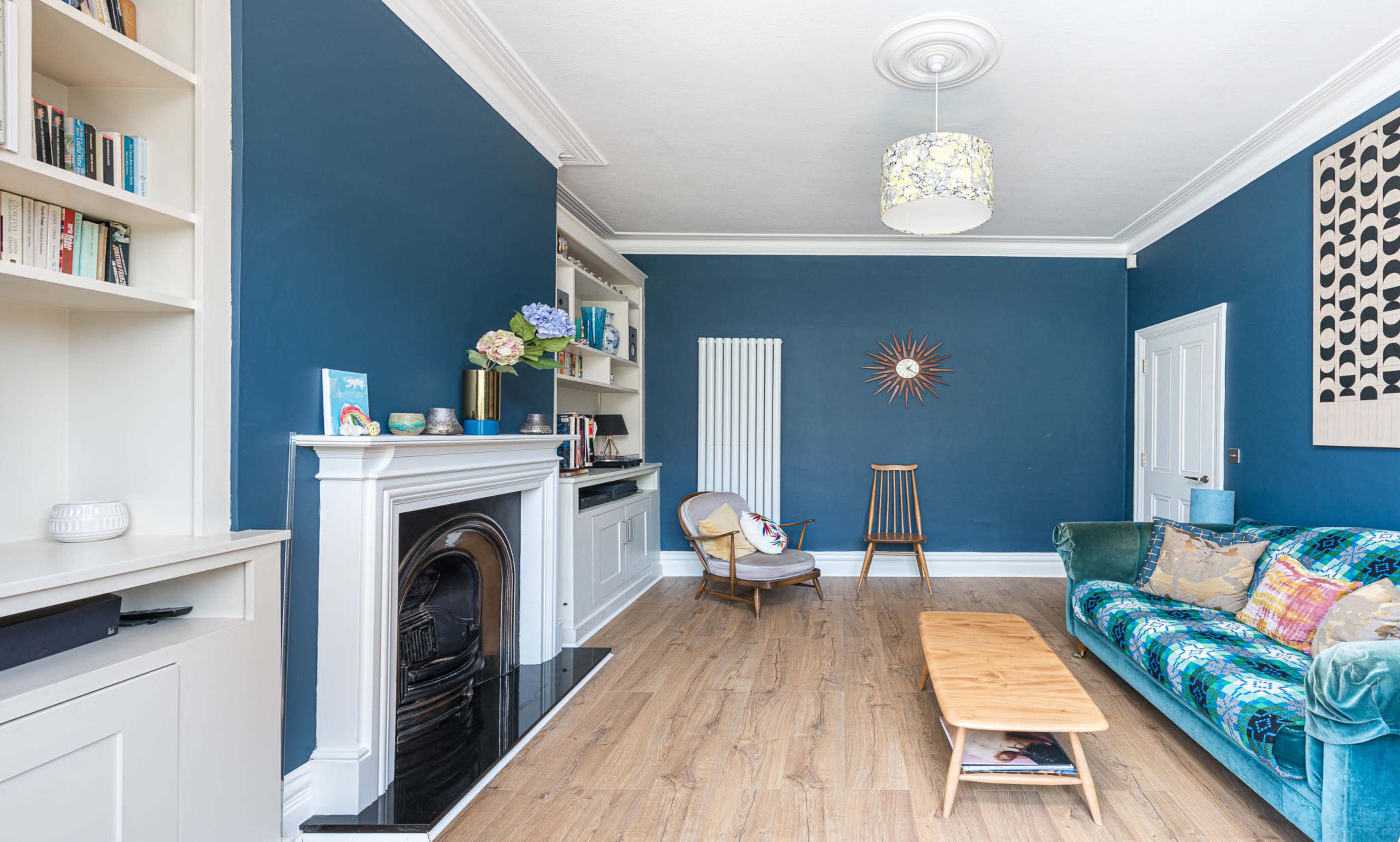 The living room features a blue accent wall, a white mantel with a decorative fireplace, a wooden coffee table, and a plush sofa adorned with colorful cushions.