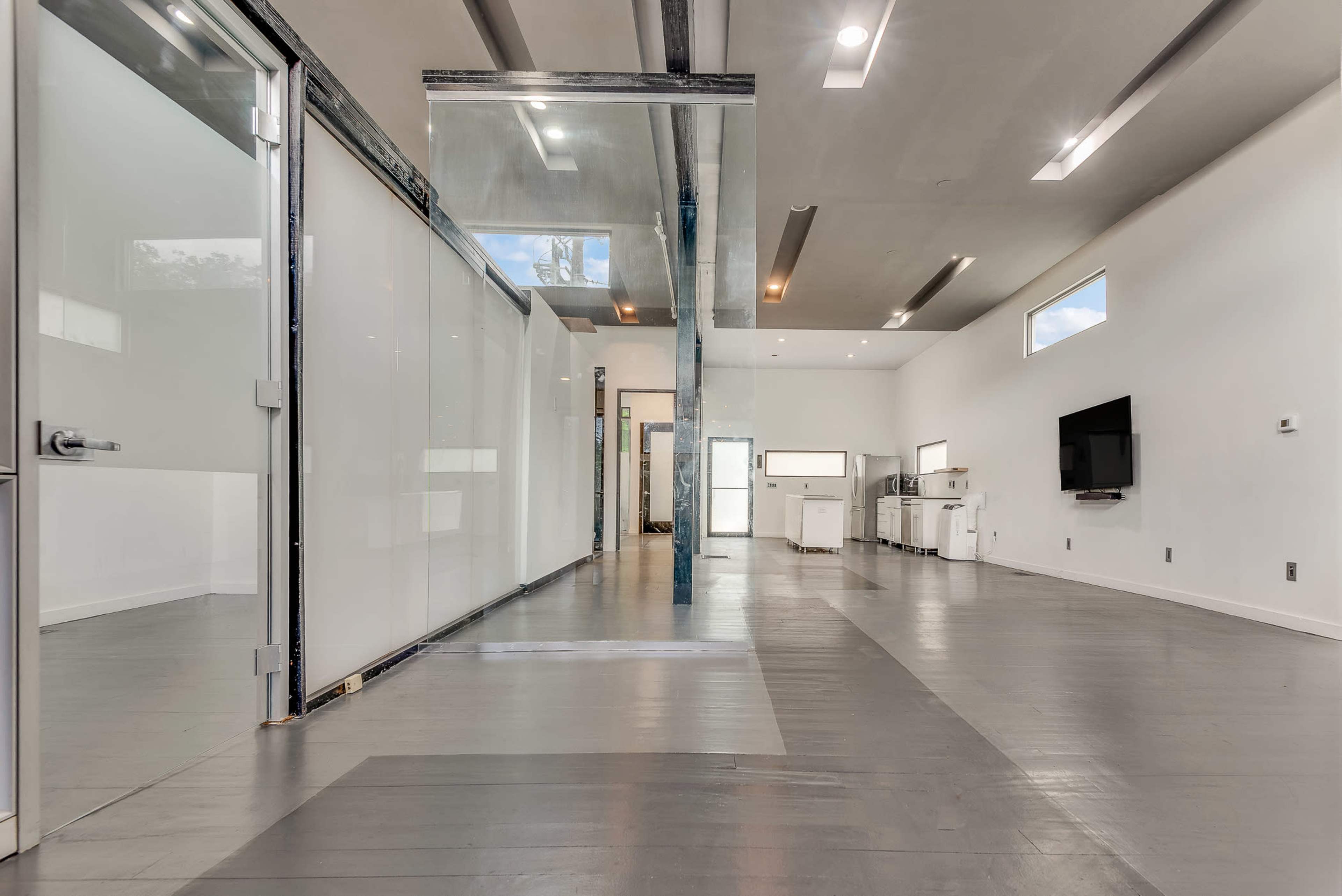 The image shows a spacious, modern interior with a combination of glass and open walls, featuring light-colored flooring and minimal furniture.