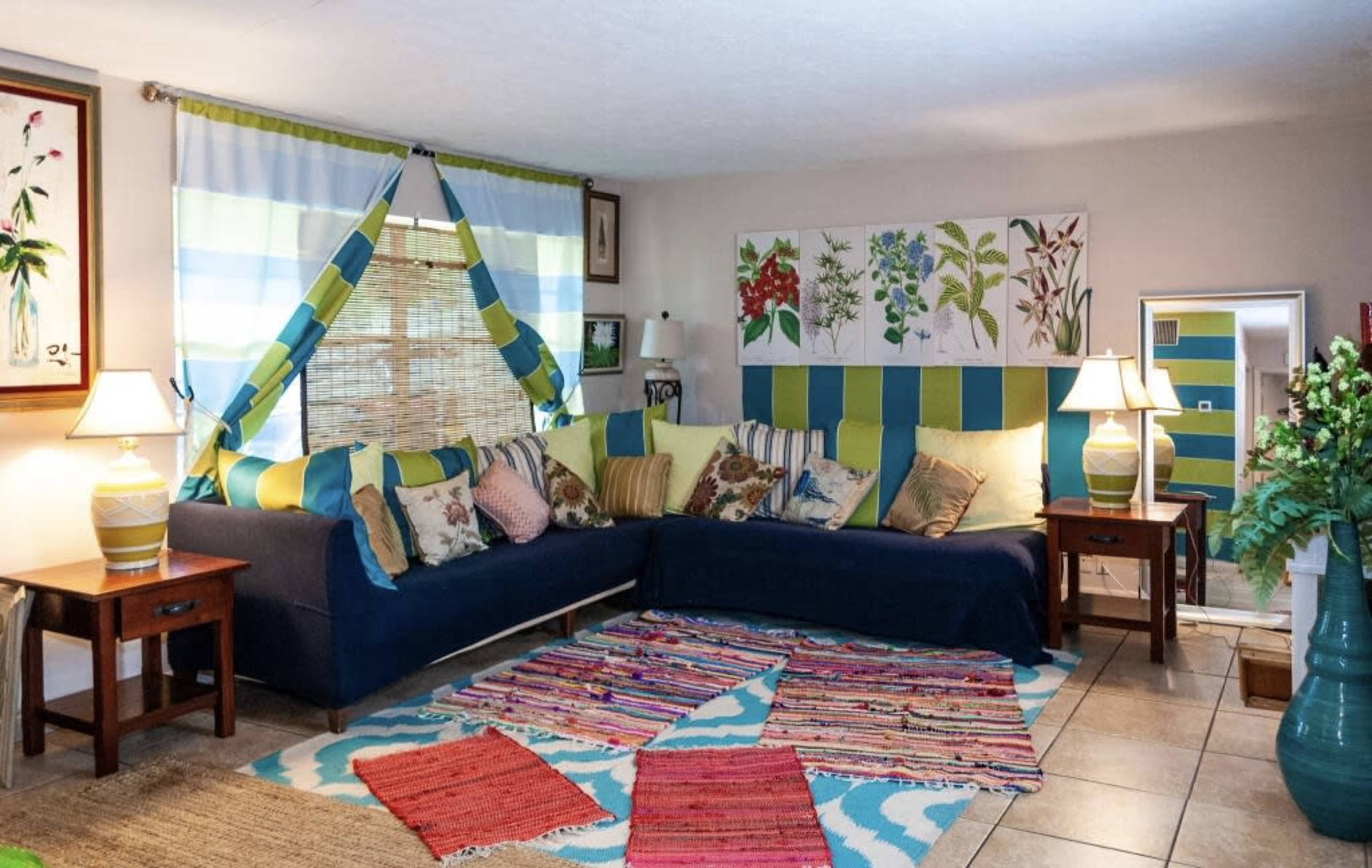 A living room features a sectional couch with colorful cushions, a patterned rug, and two lamps on wooden side tables.