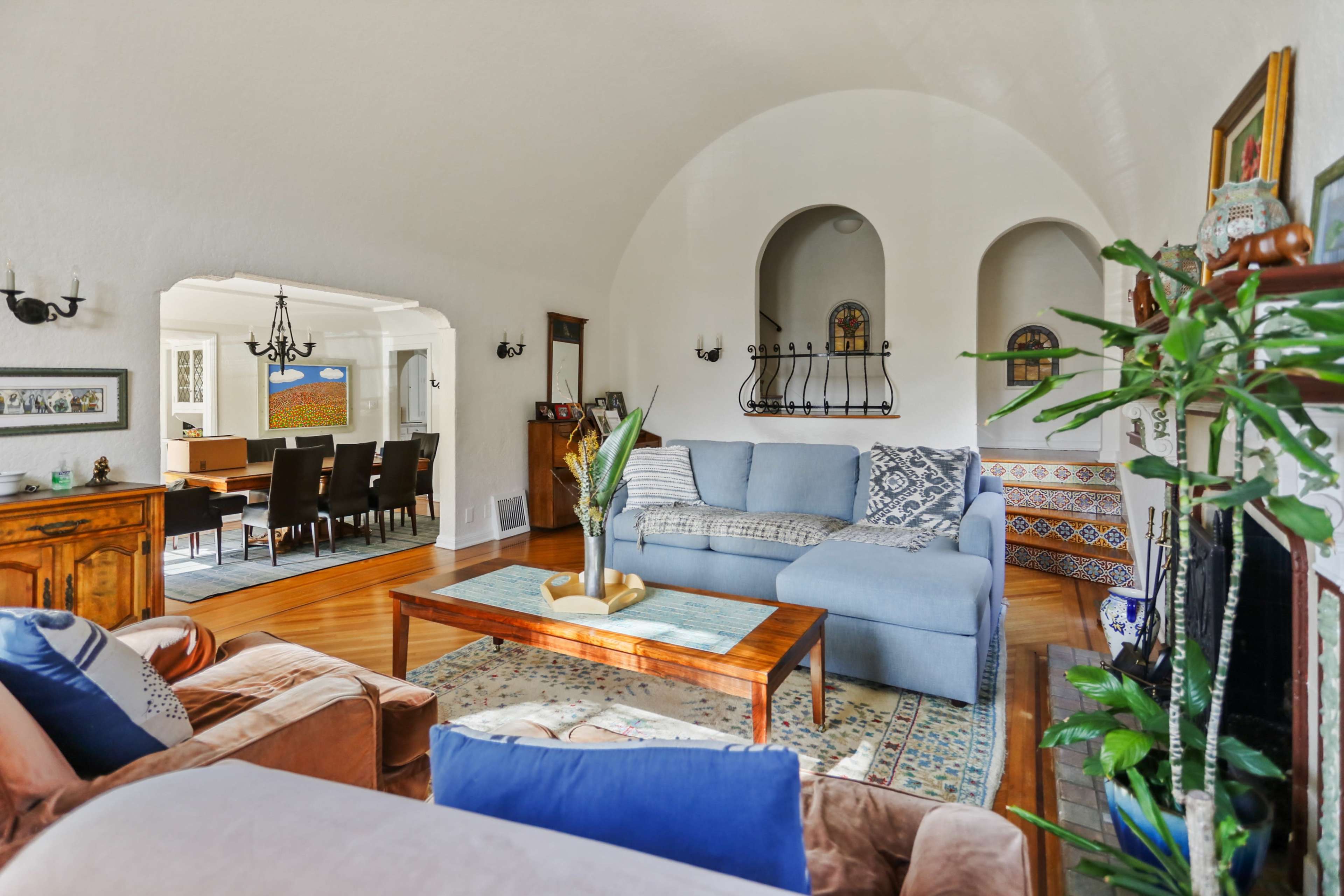 A living room with an archway leading to a dining area, featuring a blue sofa, wooden coffee table, and decorative plants.
