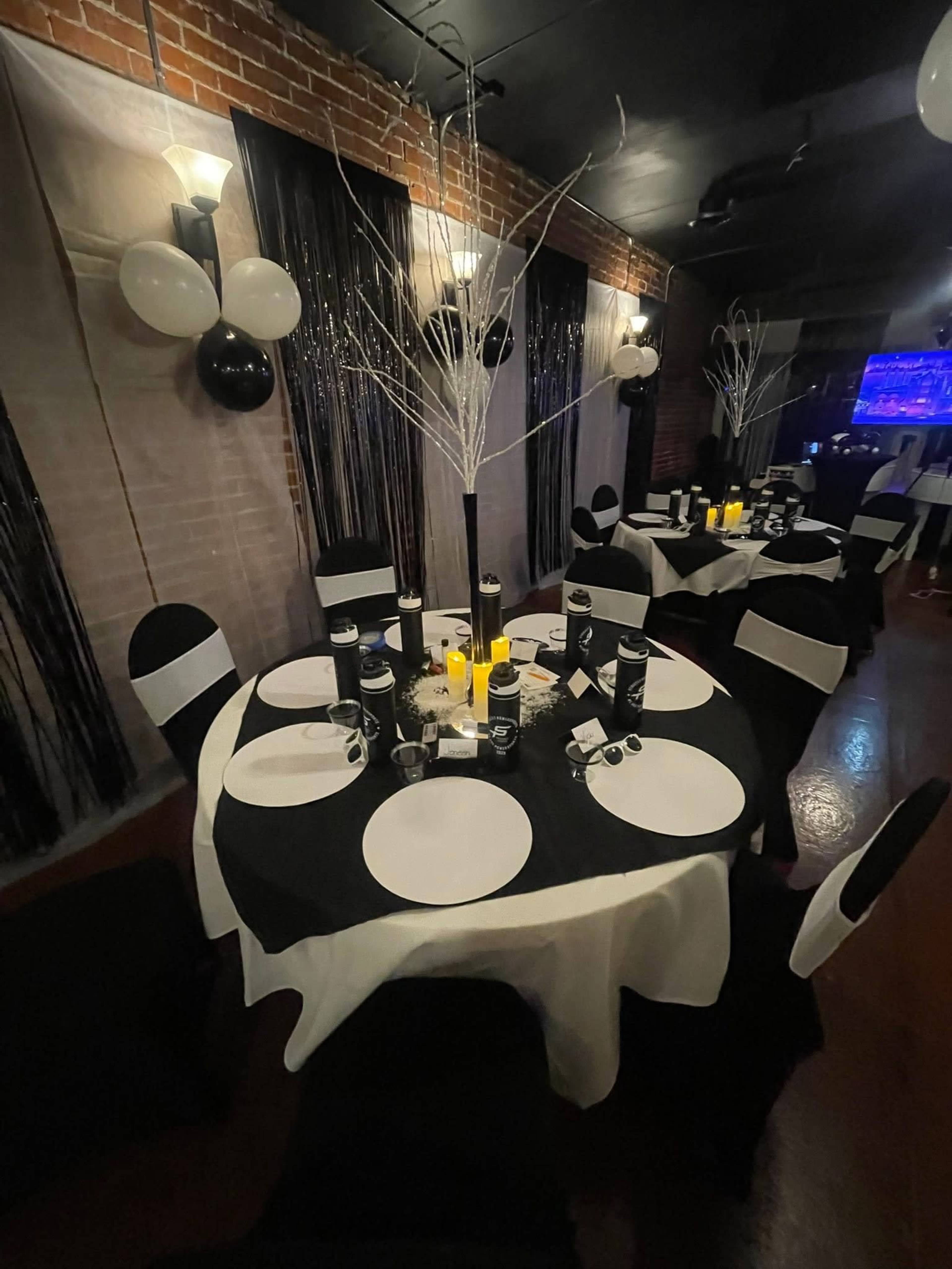 The image shows a banquet room set up with round tables covered in black and white tablecloths, decorated with candles, and accented by silver branches and balloons.