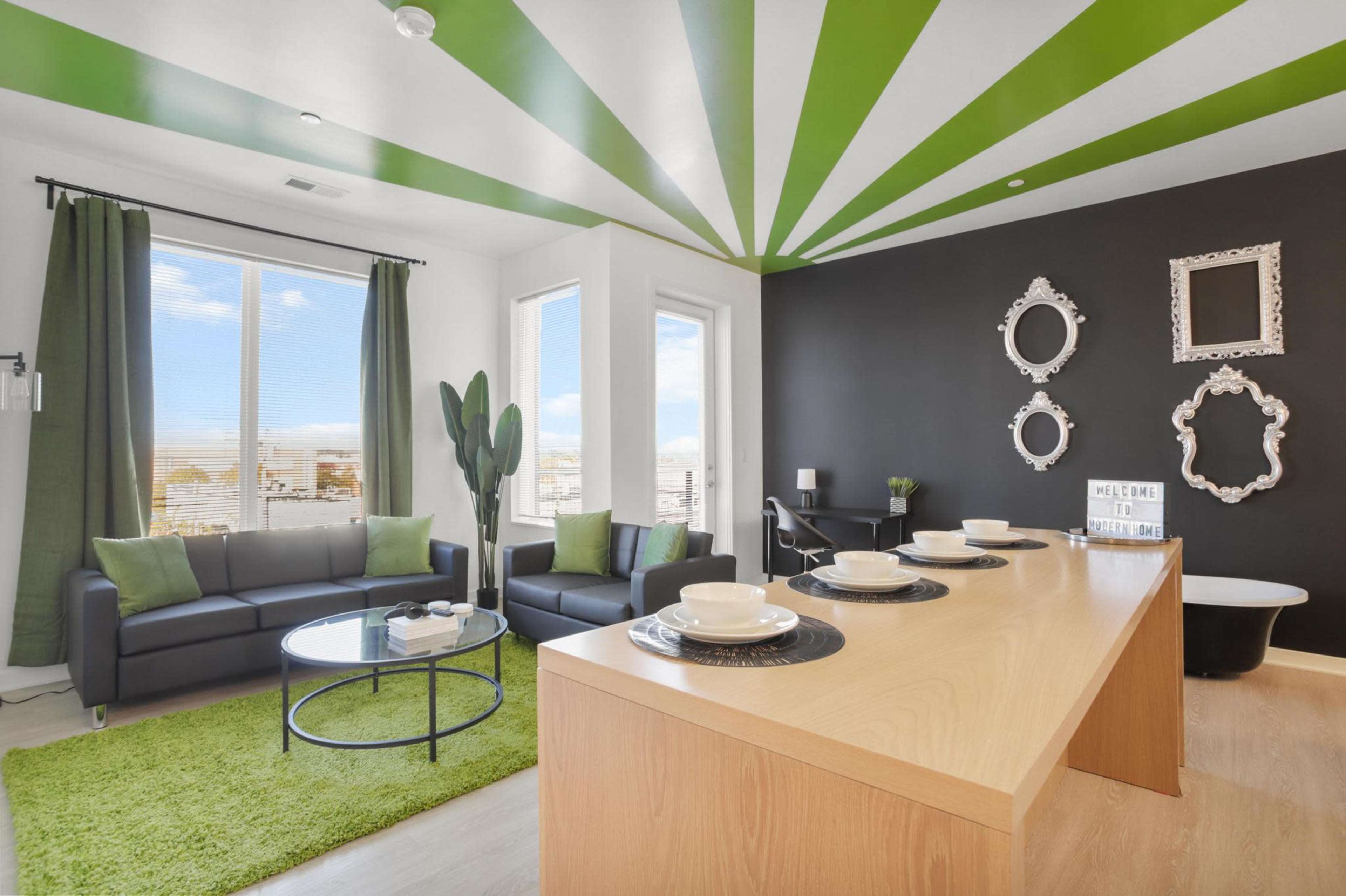 The image shows a modern living room with a striped green and white ceiling, a black accent wall, potted plants, and a wooden dining table set with plates.