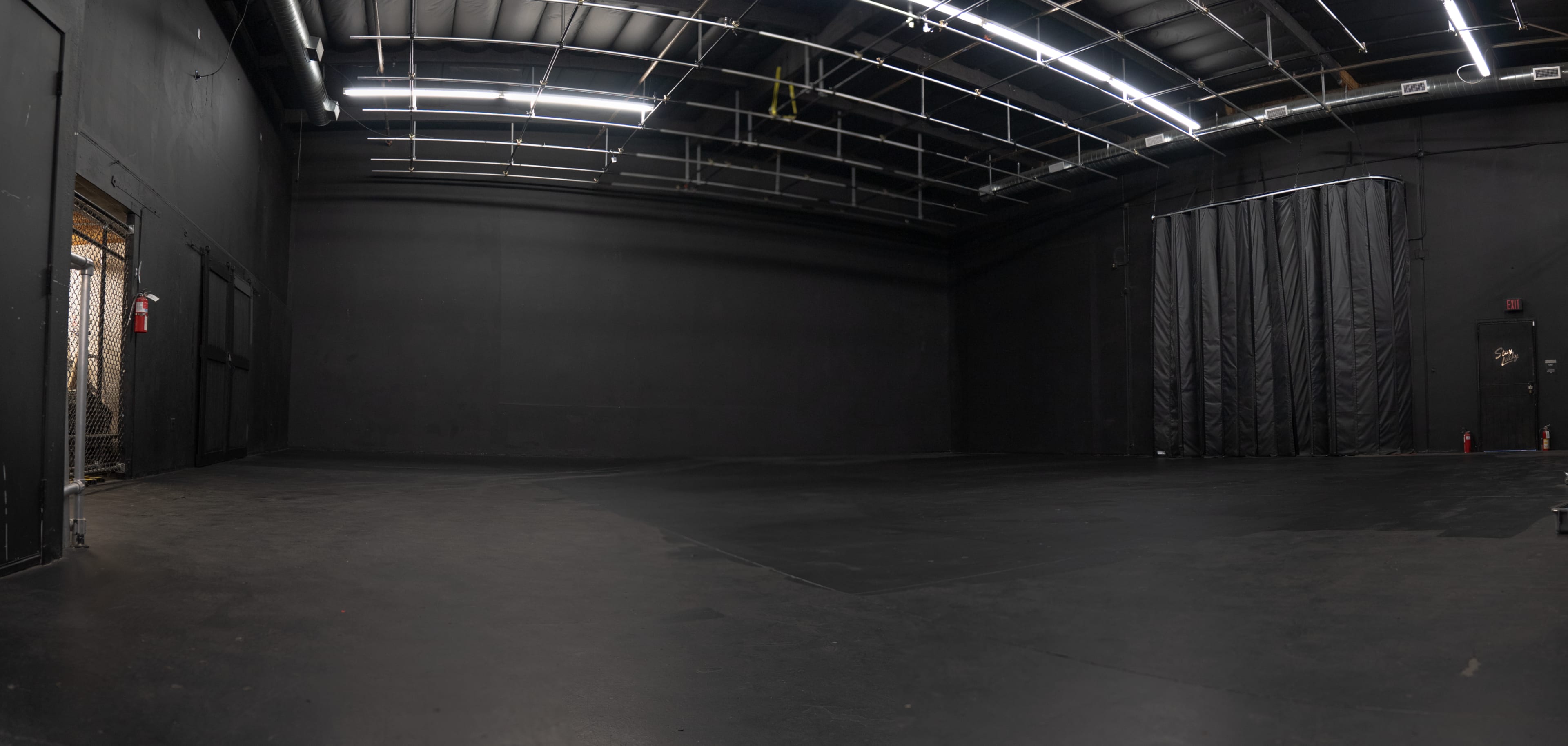The image shows a large, empty black-walled studio space with a dark floor and minimal lighting.
