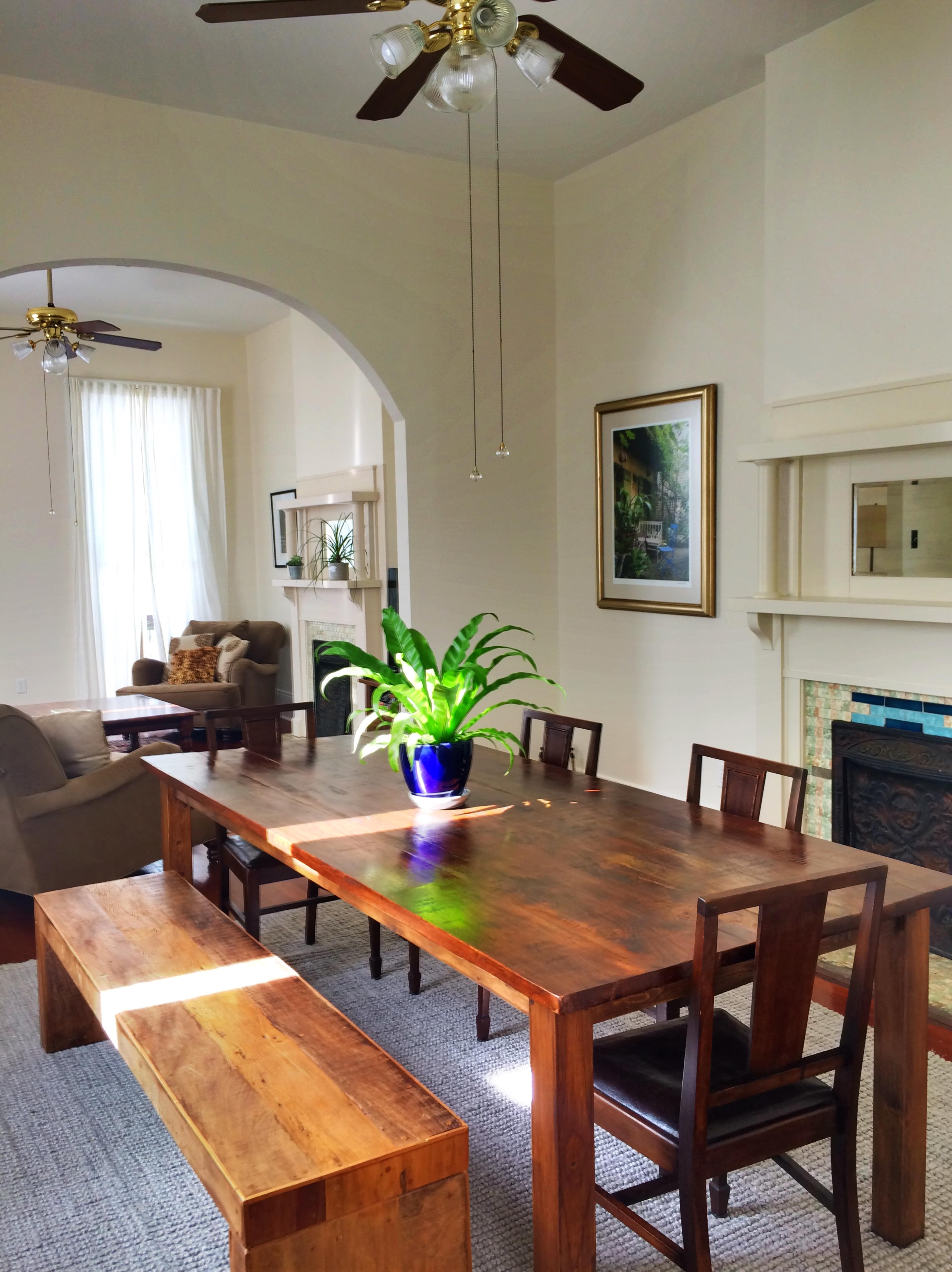 The image shows a bright dining area featuring a wooden table surrounded by chairs and a long bench, with a plant centerpiece and an archway leading to a living space.