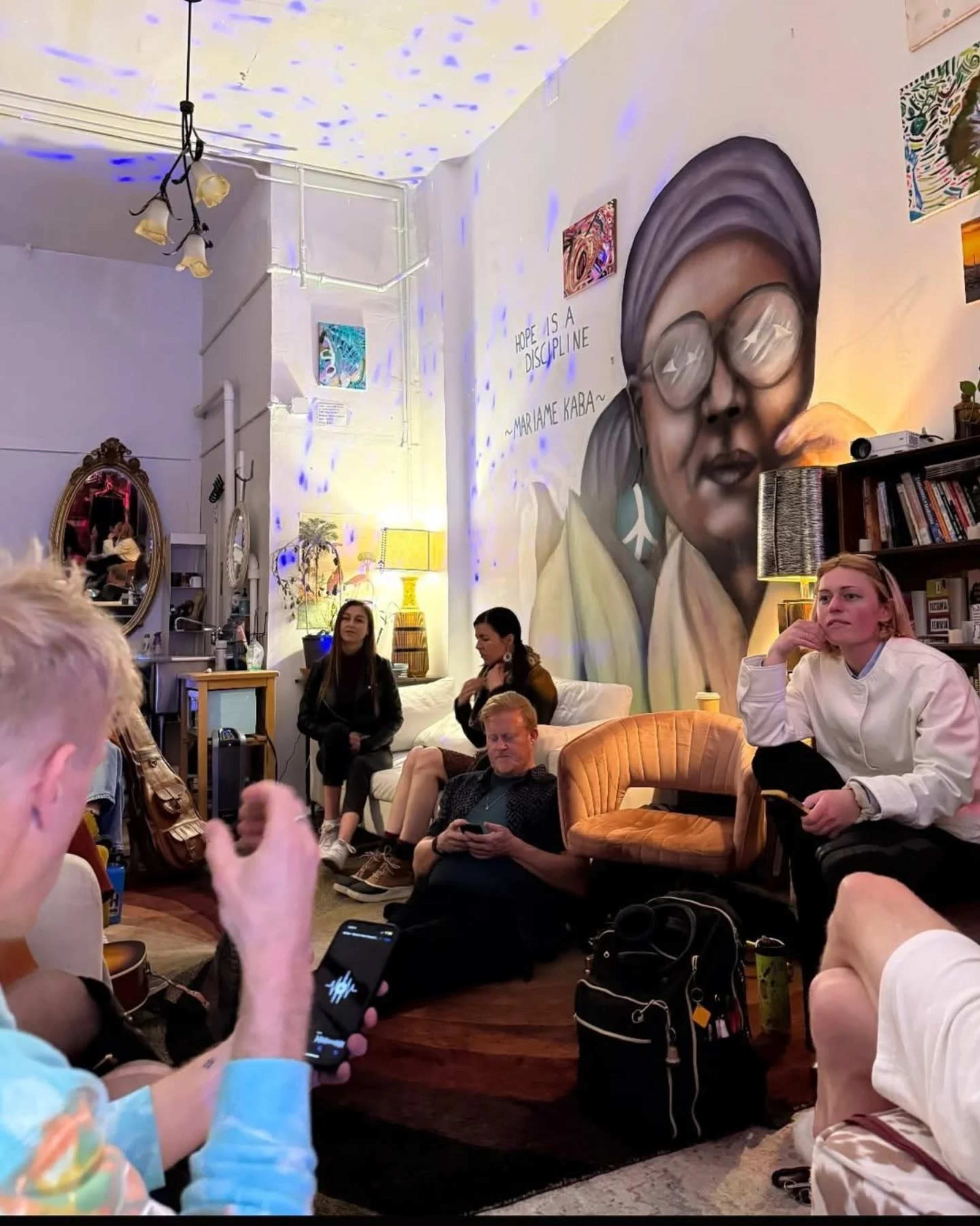 A group of people sits in a brightly lit room adorned with colorful artwork, while one person gestures with a phone in hand.