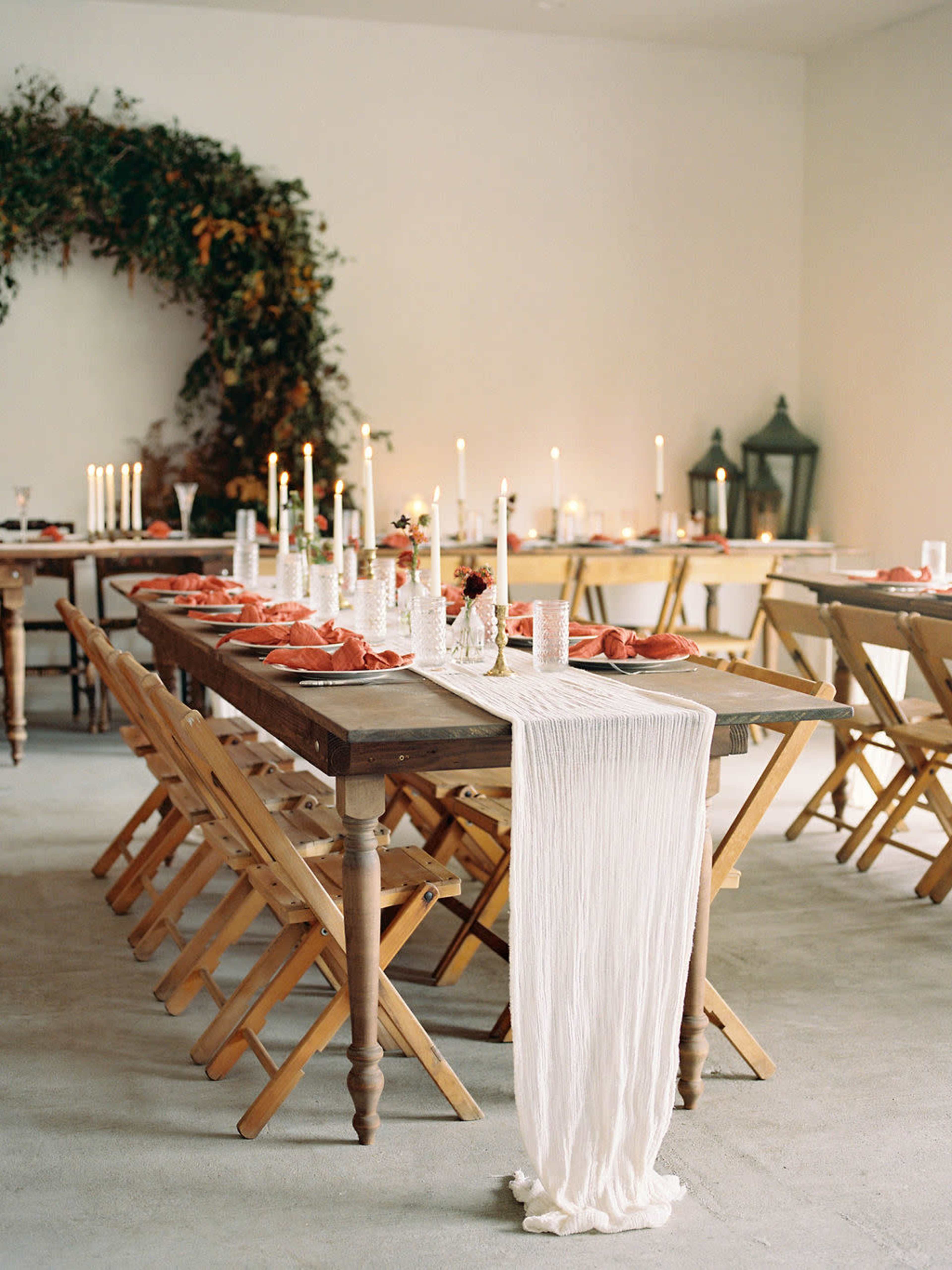 A long wooden table is set with plates and candles, surrounded by folding chairs, with greenery arching in the background.