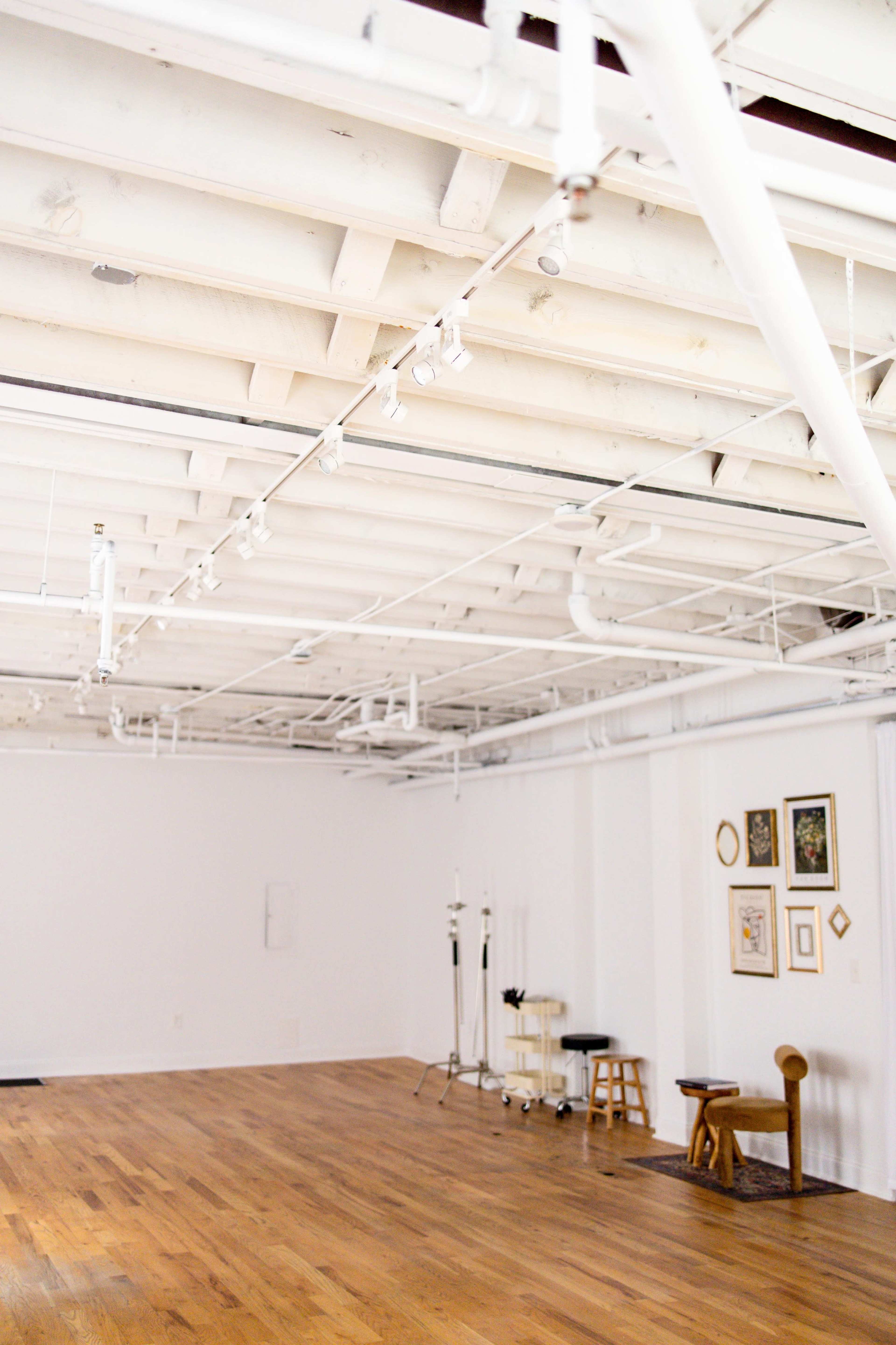 An empty, modern studio space with wooden flooring and white walls, featuring exposed ceiling beams and track lighting.