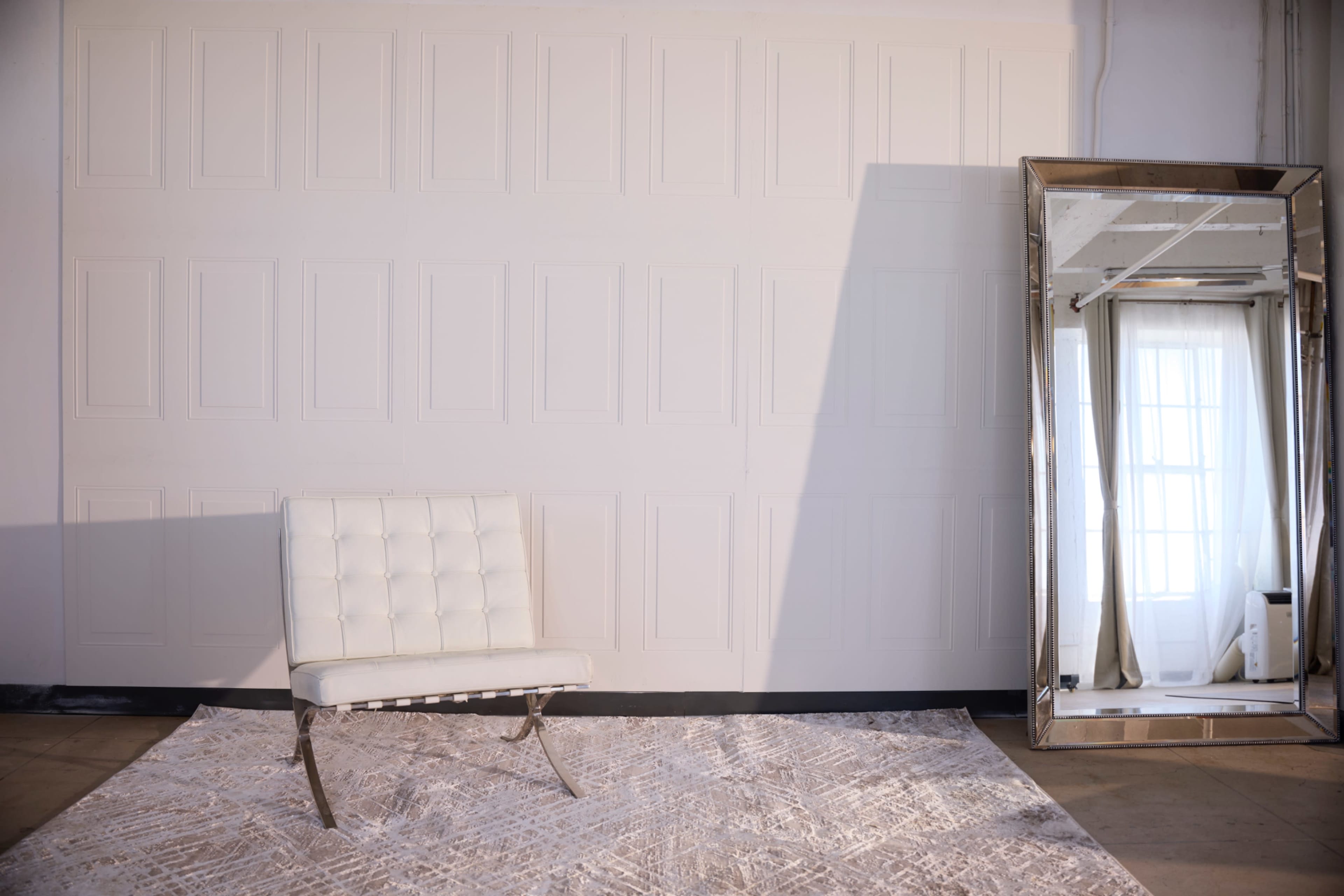 A white tufted chair is placed on a patterned rug next to a large mirror reflecting a window.