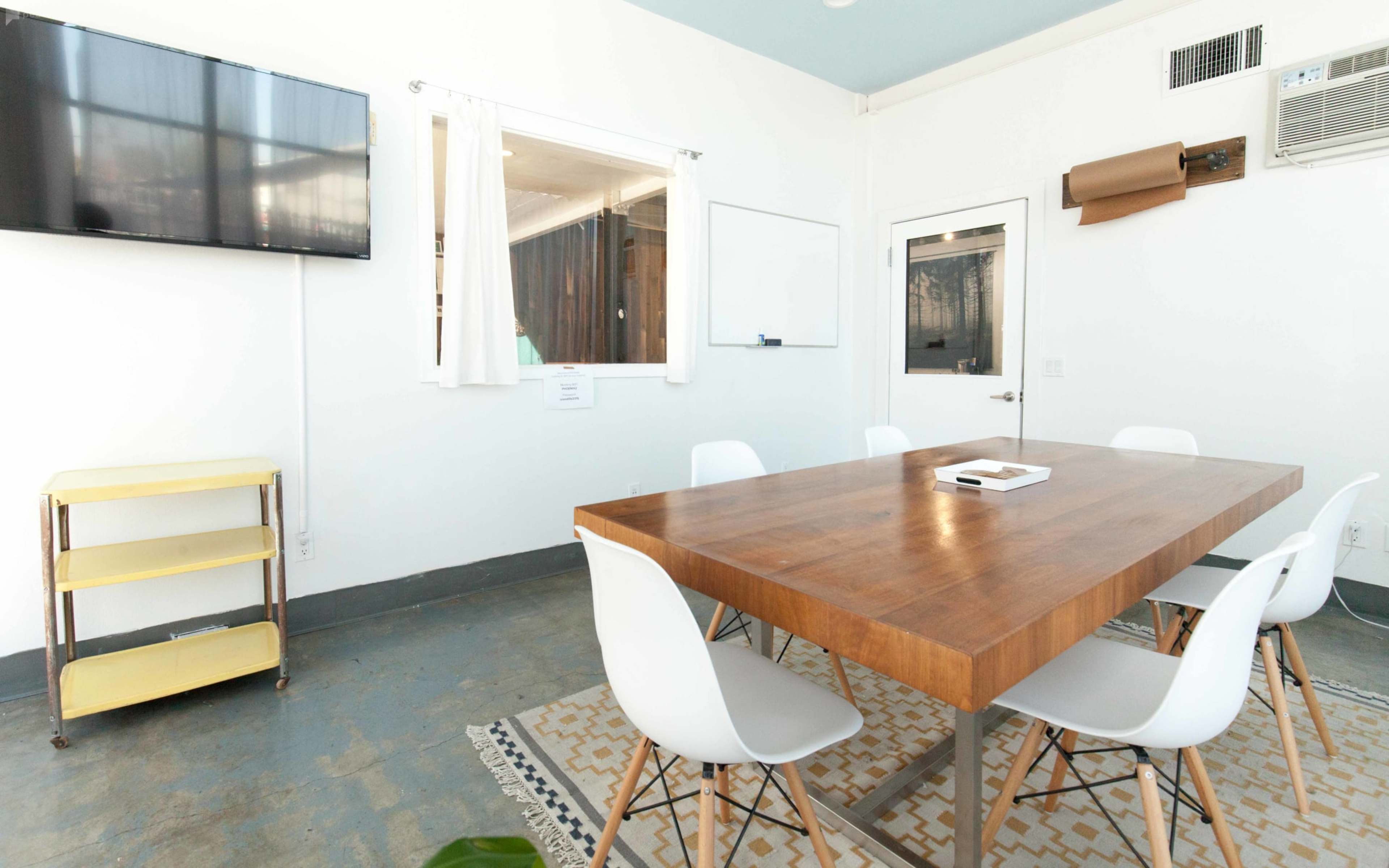A conference room features a wooden table surrounded by white chairs, a television mounted on the wall, and a whiteboard.