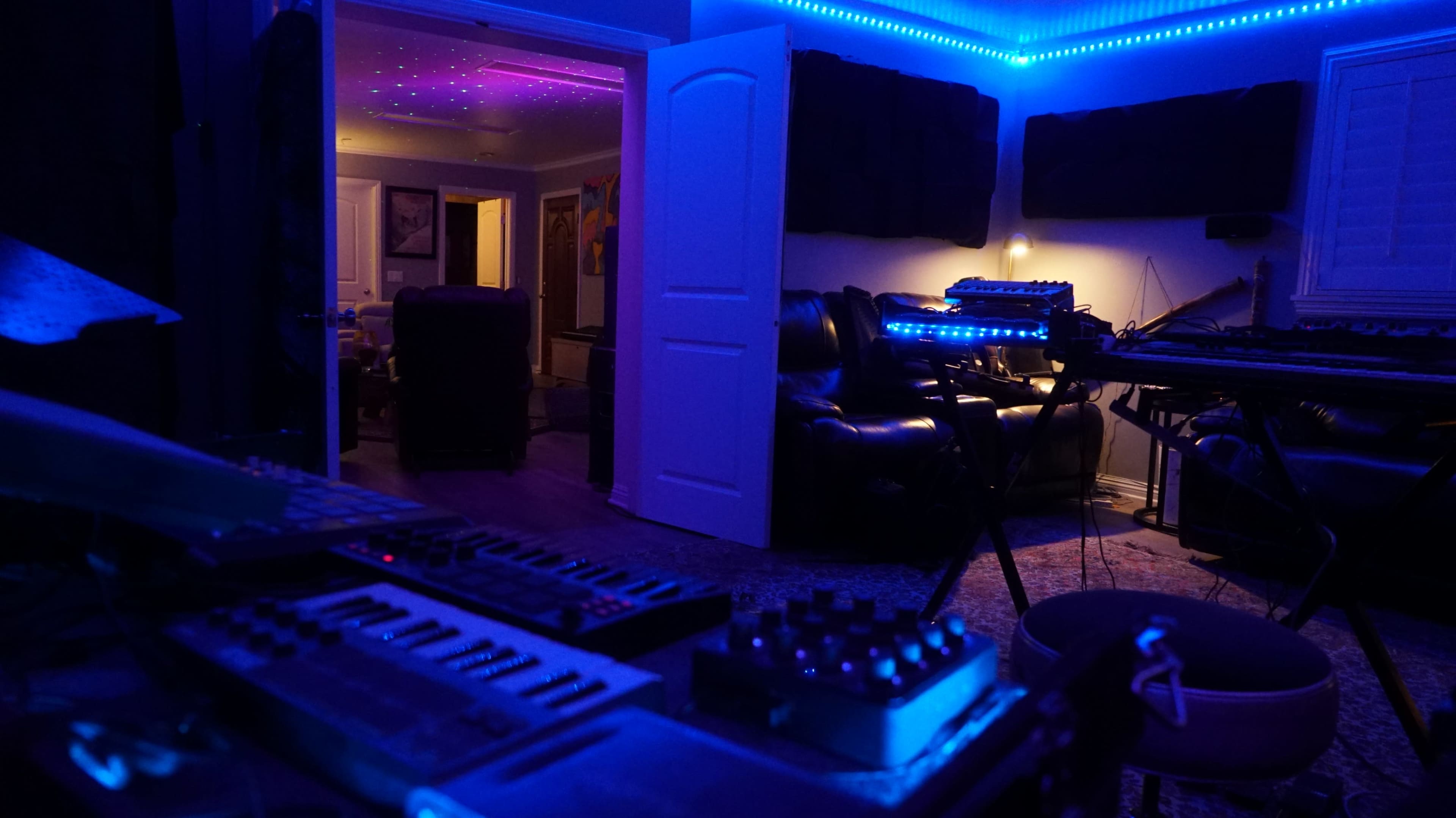 A dimly lit music studio with various synthesizers and equipment in the foreground, and a doorway leading to a different room in the background.