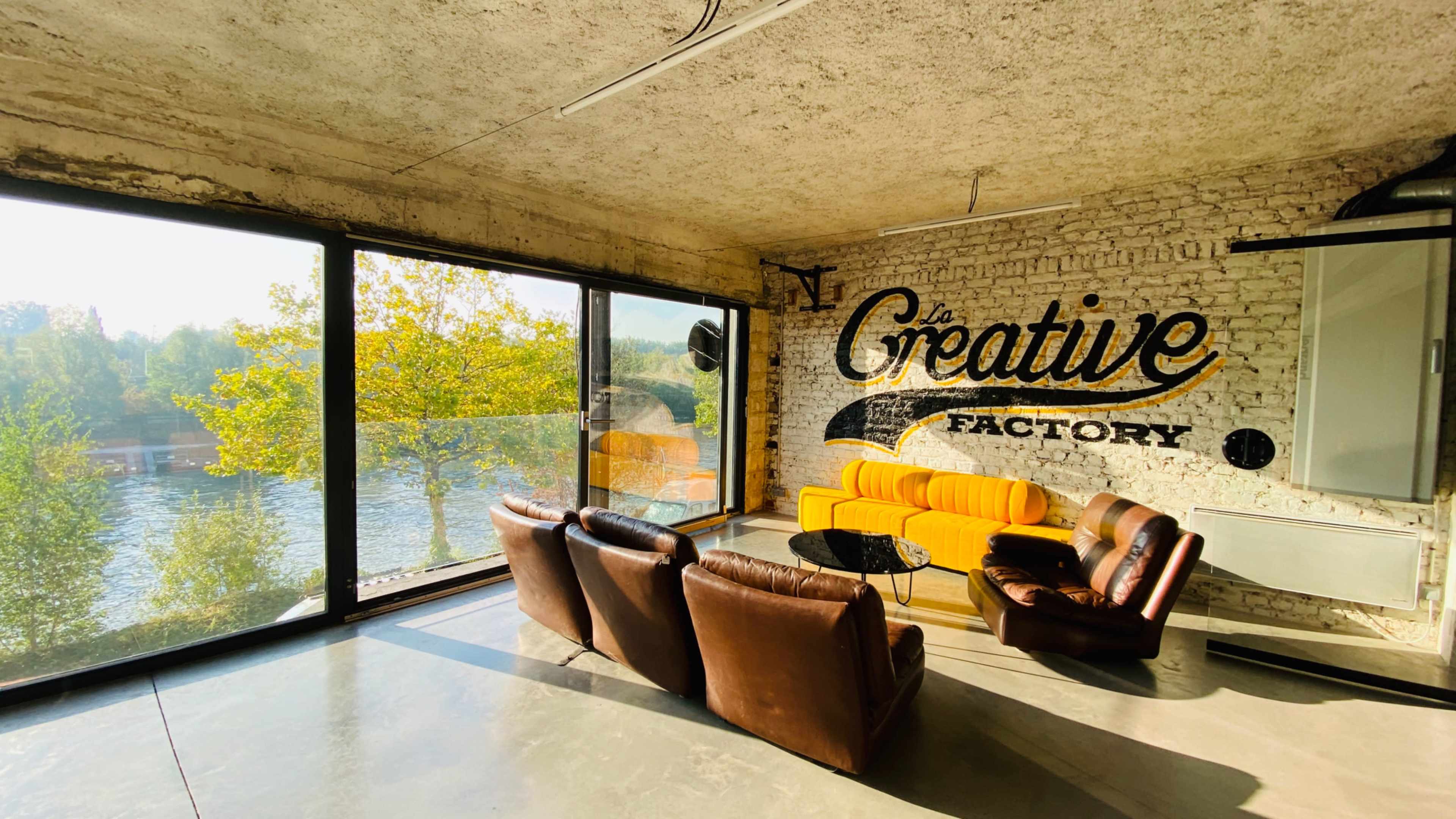 The image shows a modern interior space with large windows overlooking a body of water, featuring a distinctive wall mural that reads "Creative Factory" and a mix of leather seating arrangements.