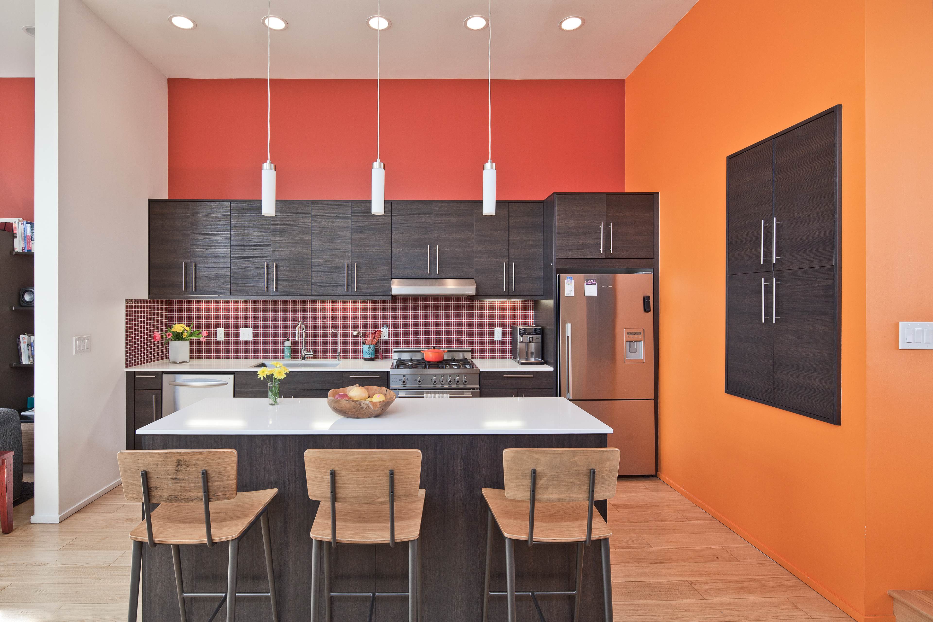 A modern kitchen features dark cabinetry, stainless steel appliances, and a bright orange accent wall with bar stools at the island.