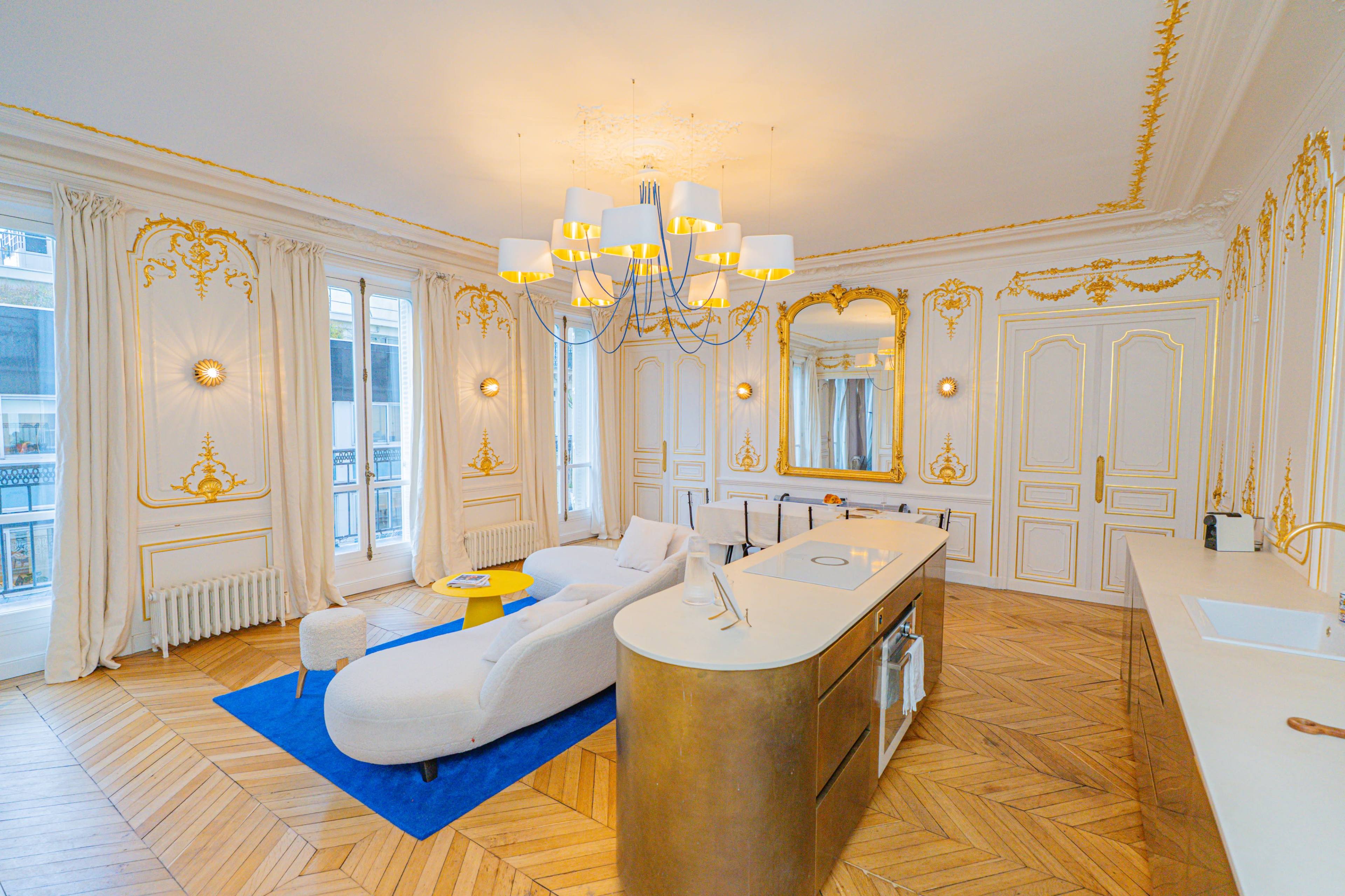 The image shows a luxurious living room featuring ornate golden details, a modern white sofa, and a central island with a sink in an elegant, well-lit space.