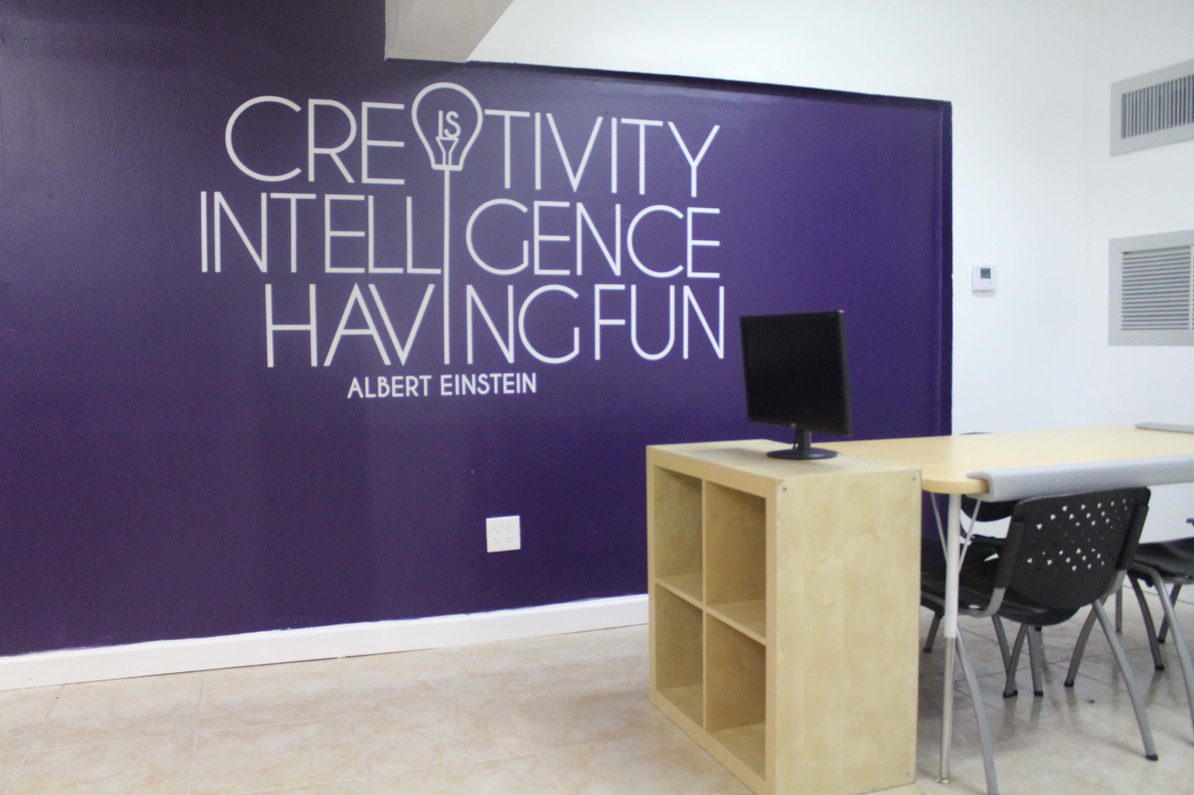 The image shows a white and purple room with a table, chairs, and a computer monitor, featuring a wall with a quote attributed to Albert Einstein about creativity, intelligence, and having fun.