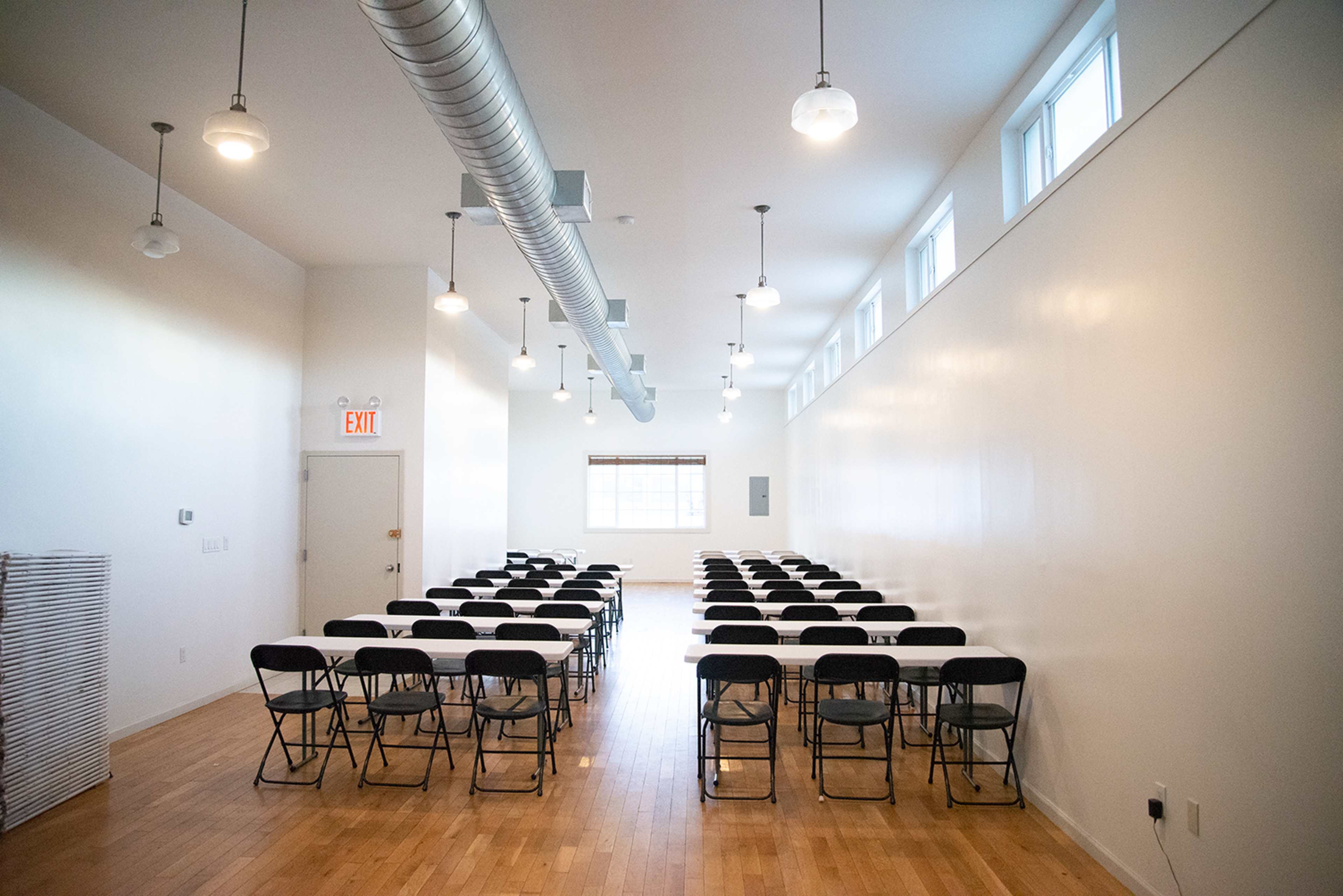 A row of black folding chairs is arranged in a rectangular formation in a bright, sparsely decorated room with large windows and overhead lights.