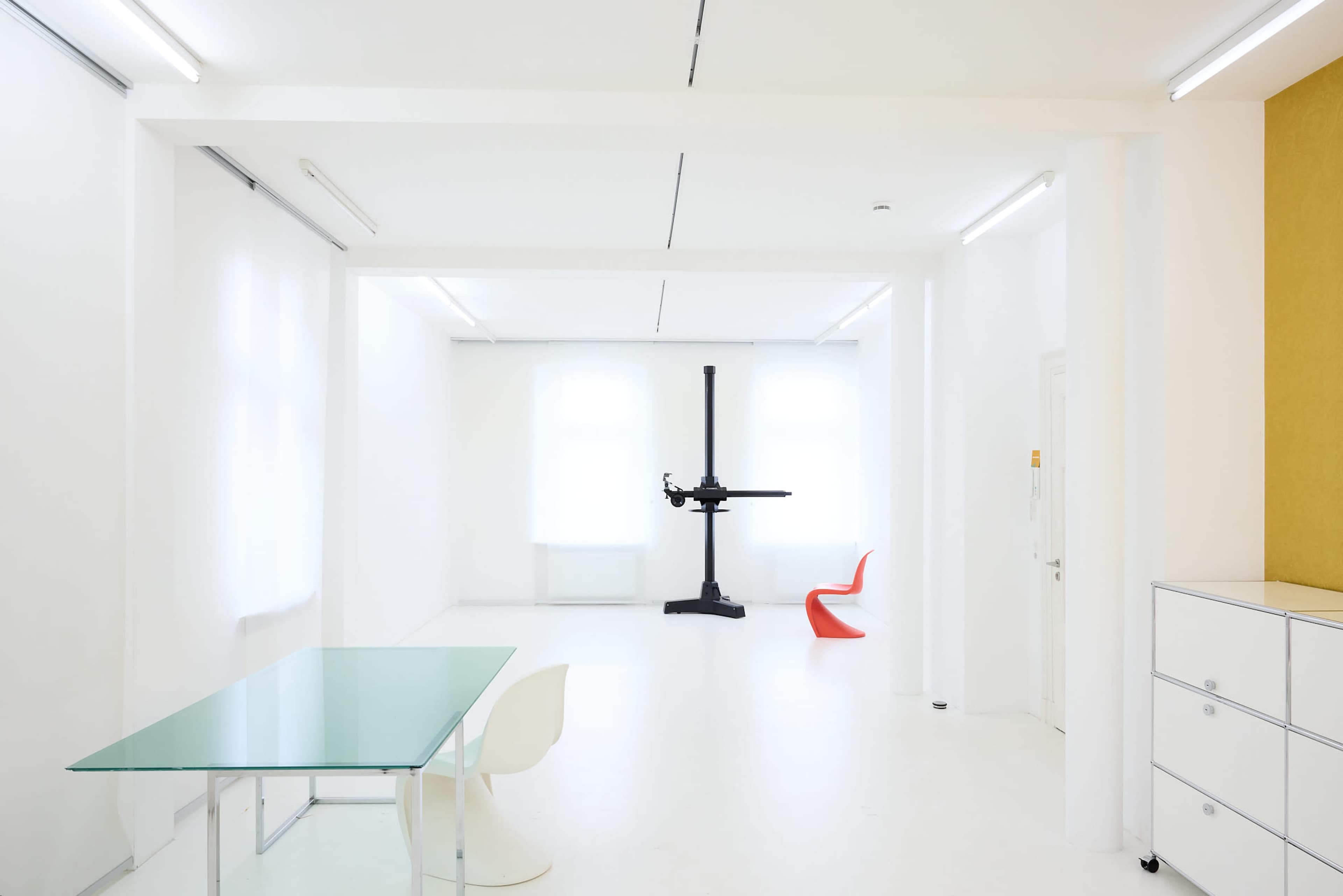 The scene features a minimalist white room with large windows, a glass table, a red chair, and a large black sculptural installation.