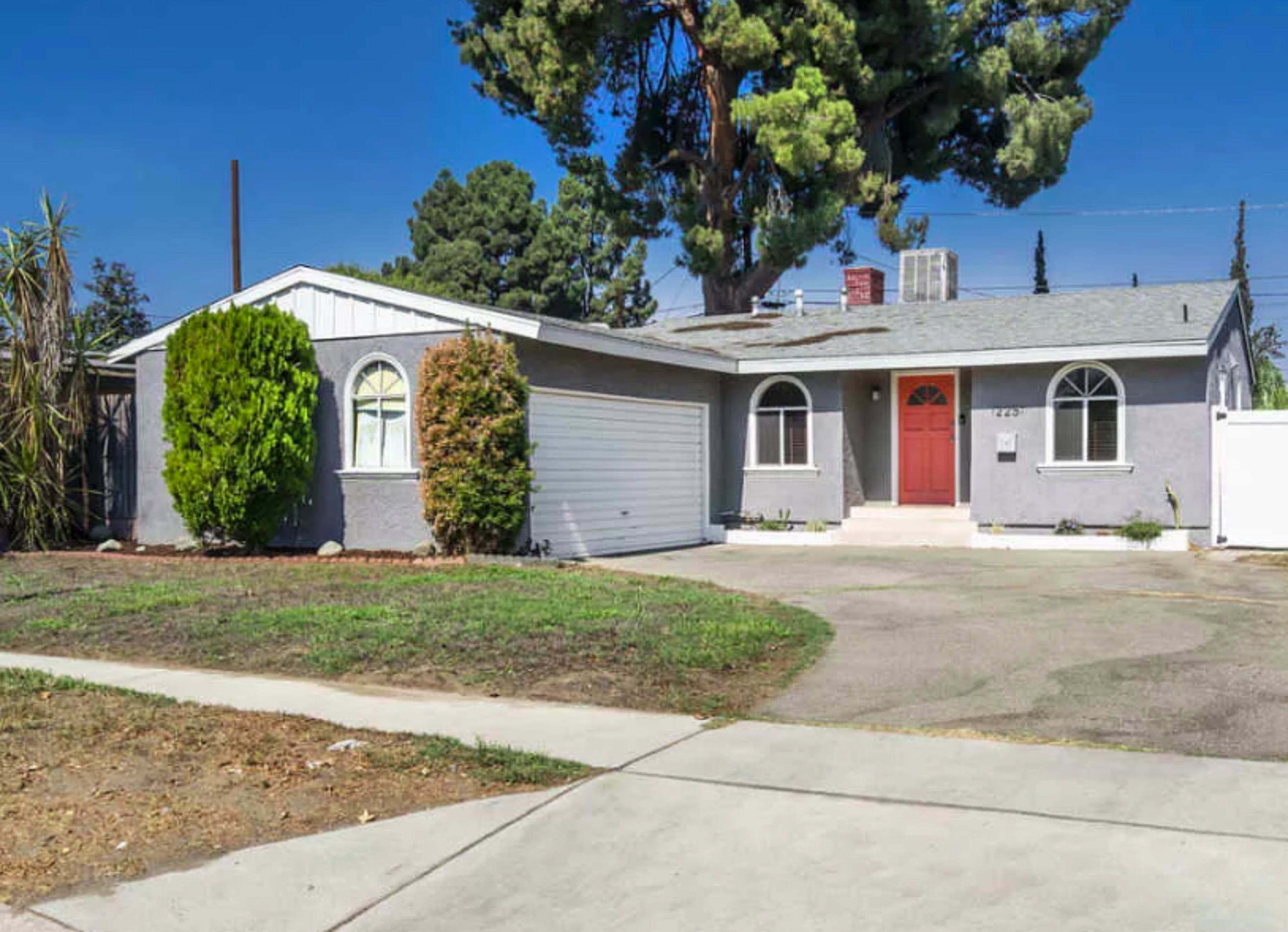 A single-story house with a gray exterior, a red front door, and a spacious driveway surrounded by a patch of grass.