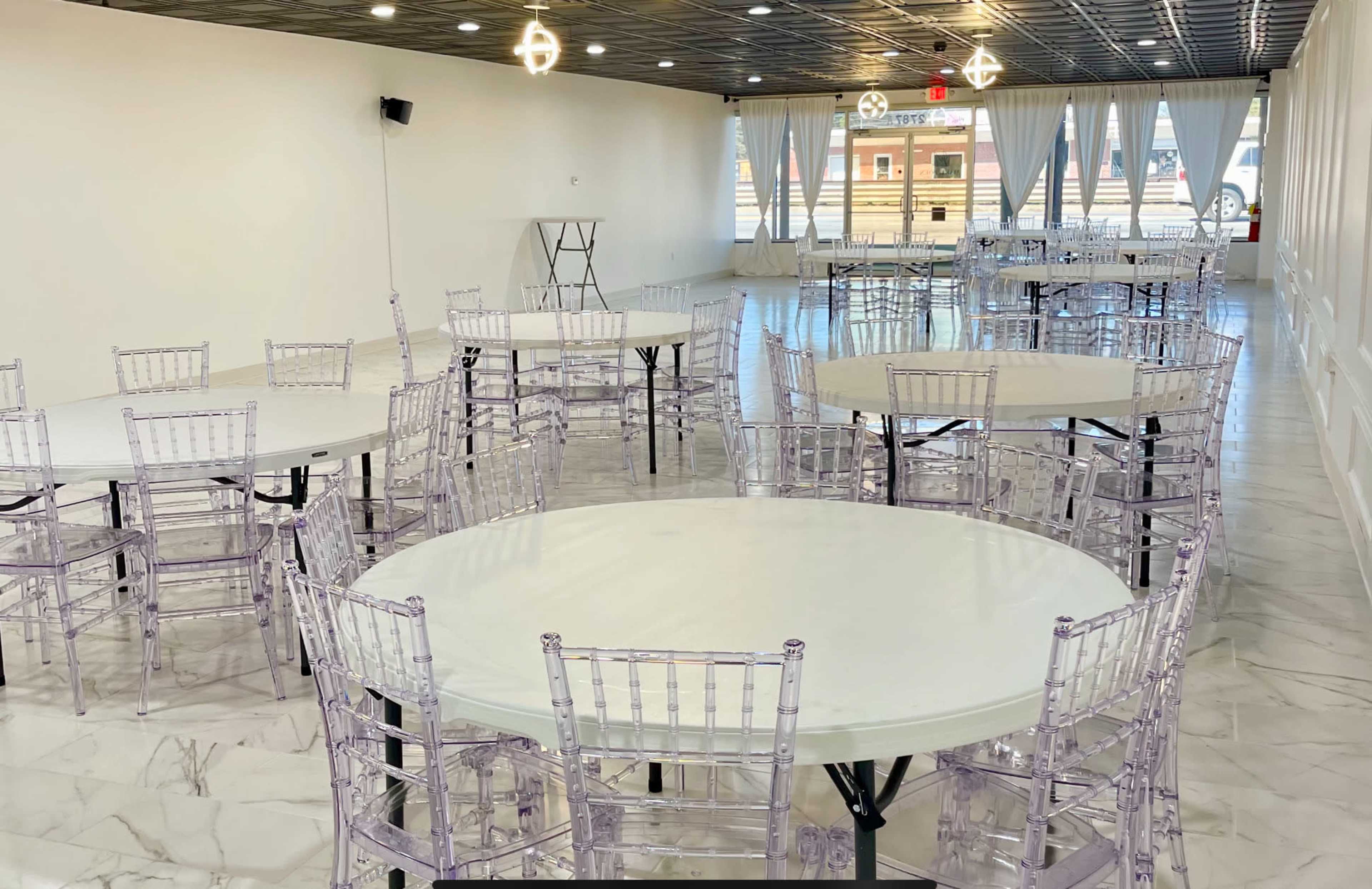A spacious event venue with several round tables surrounded by clear chiavari chairs, and large windows allowing natural light.