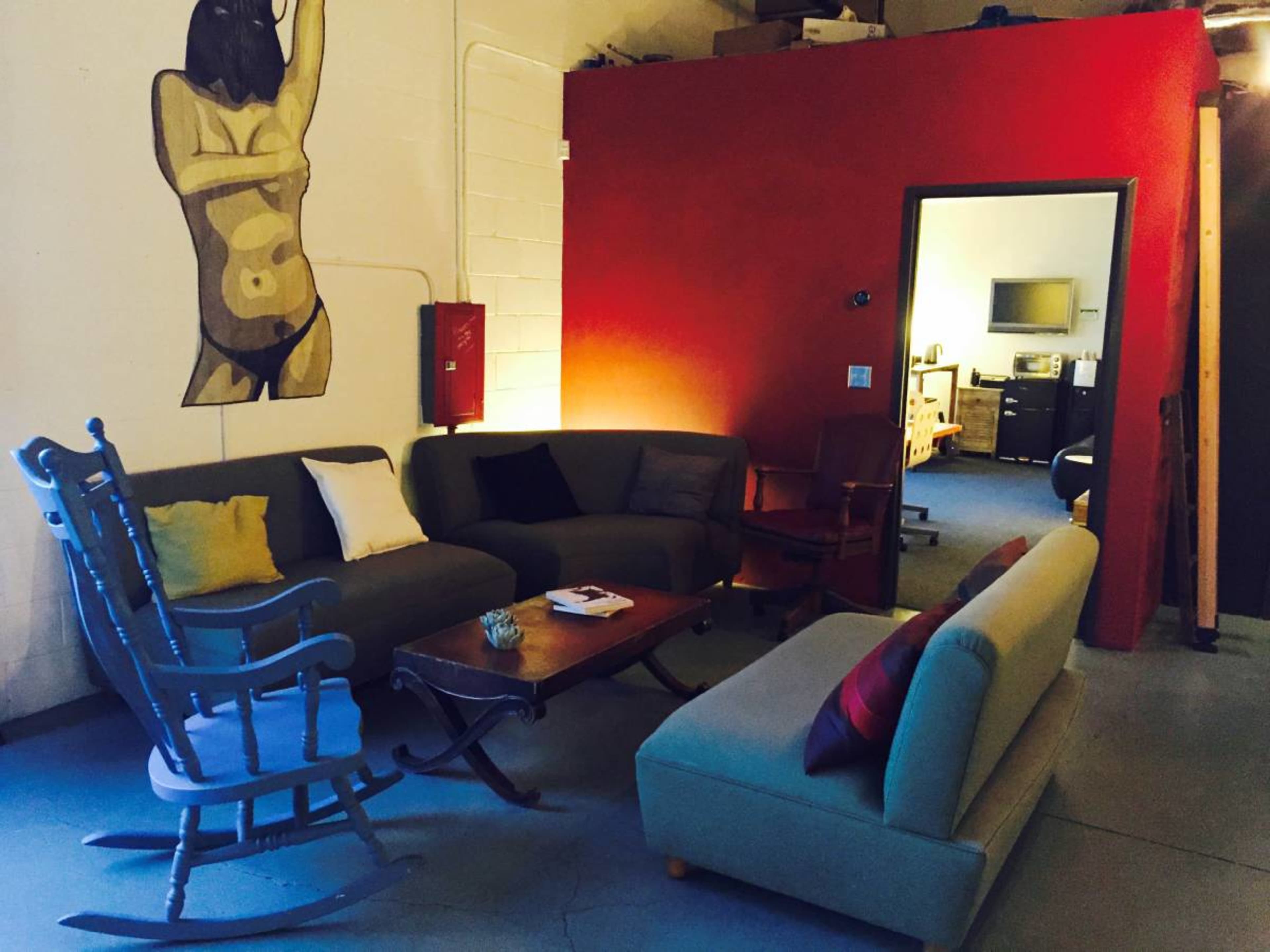 A cozy shared workspace with a red accent wall, two sofas, a rocking chair, and a view into an office area with a desk and computer.
