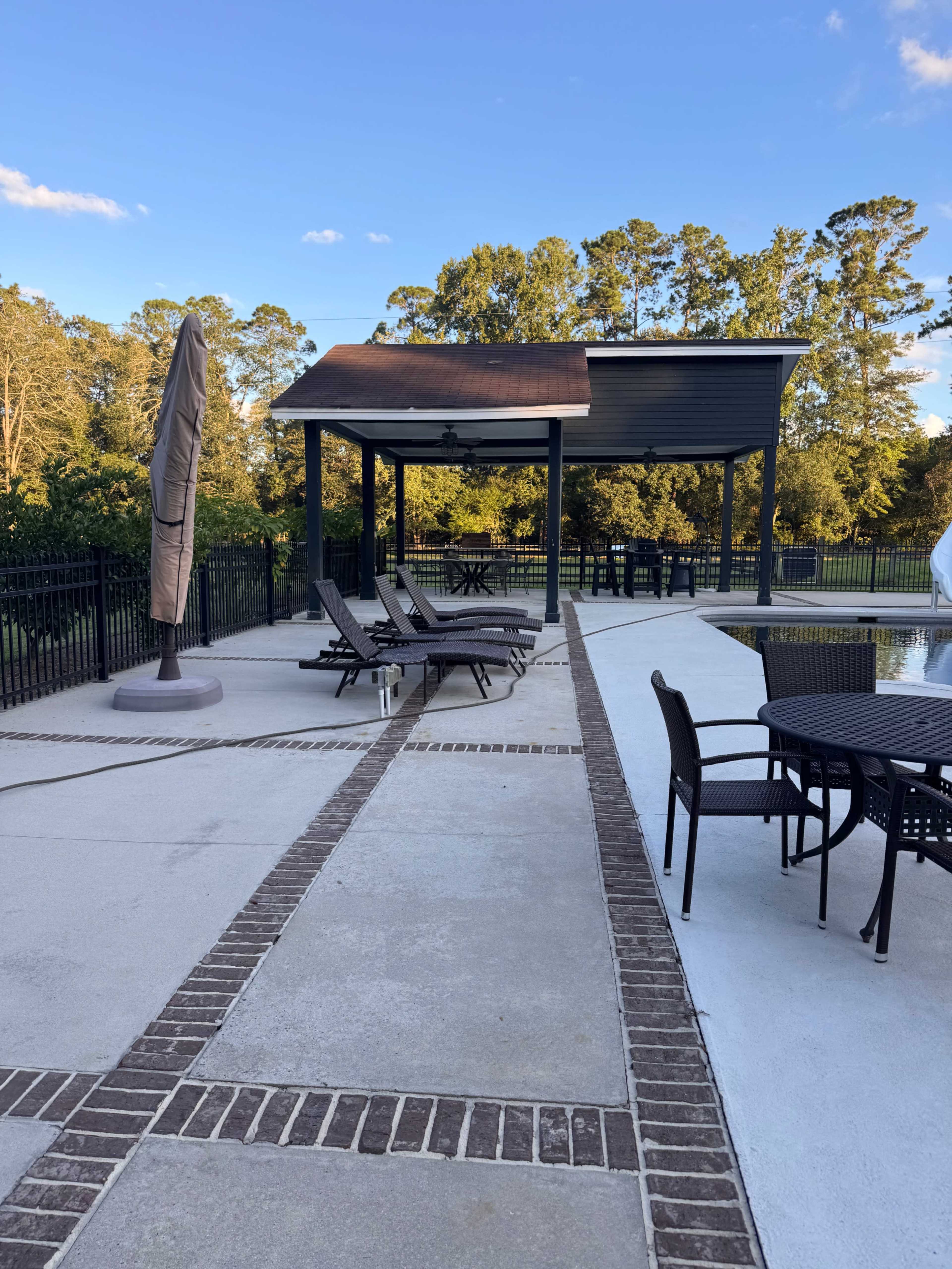 The image shows a poolside area with lounge chairs and a covered seating space surrounded by trees.