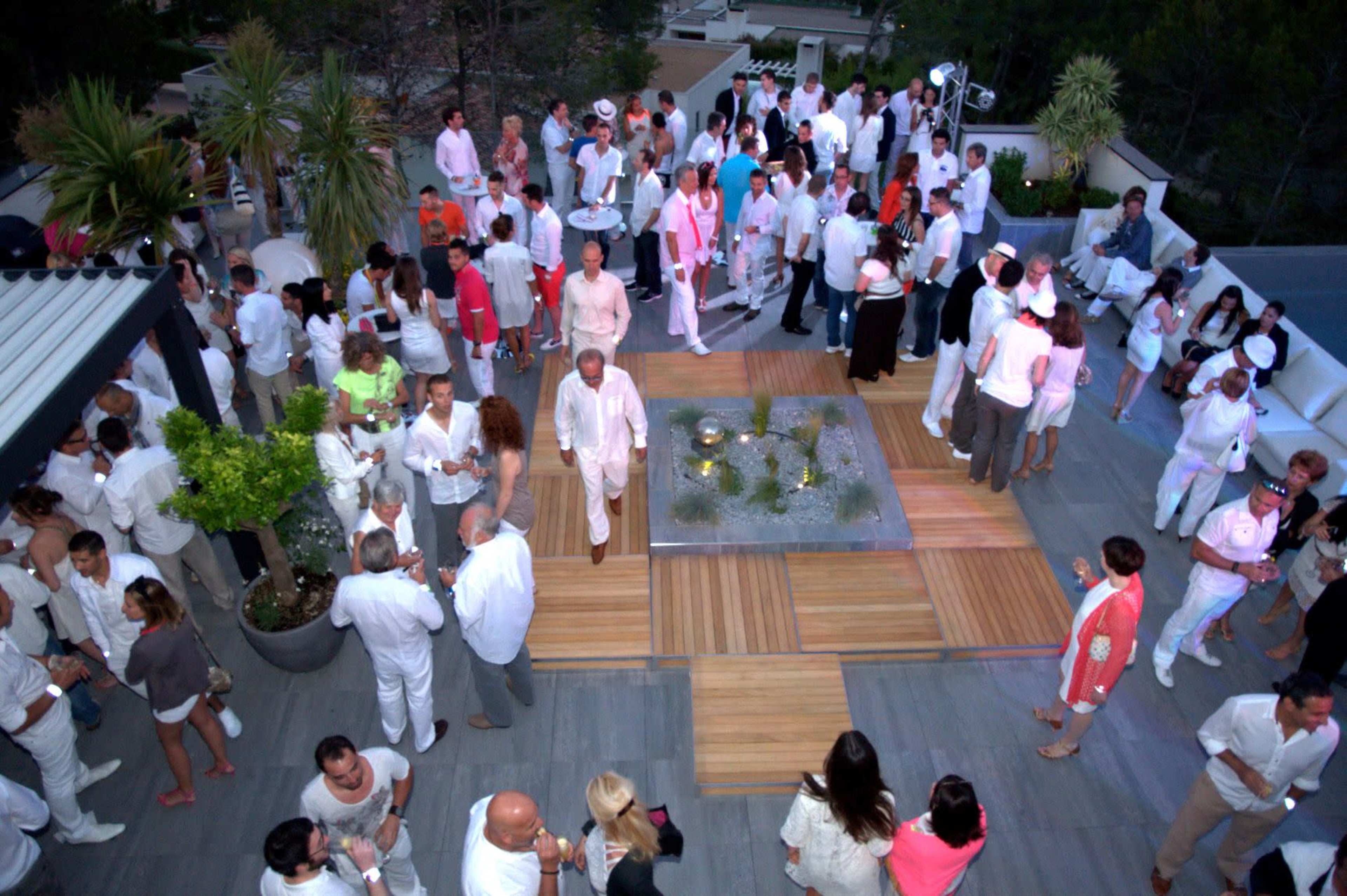 A large group of people dressed in white attire socializes on an outdoor terrace featuring wooden flooring and a glass centerpiece.