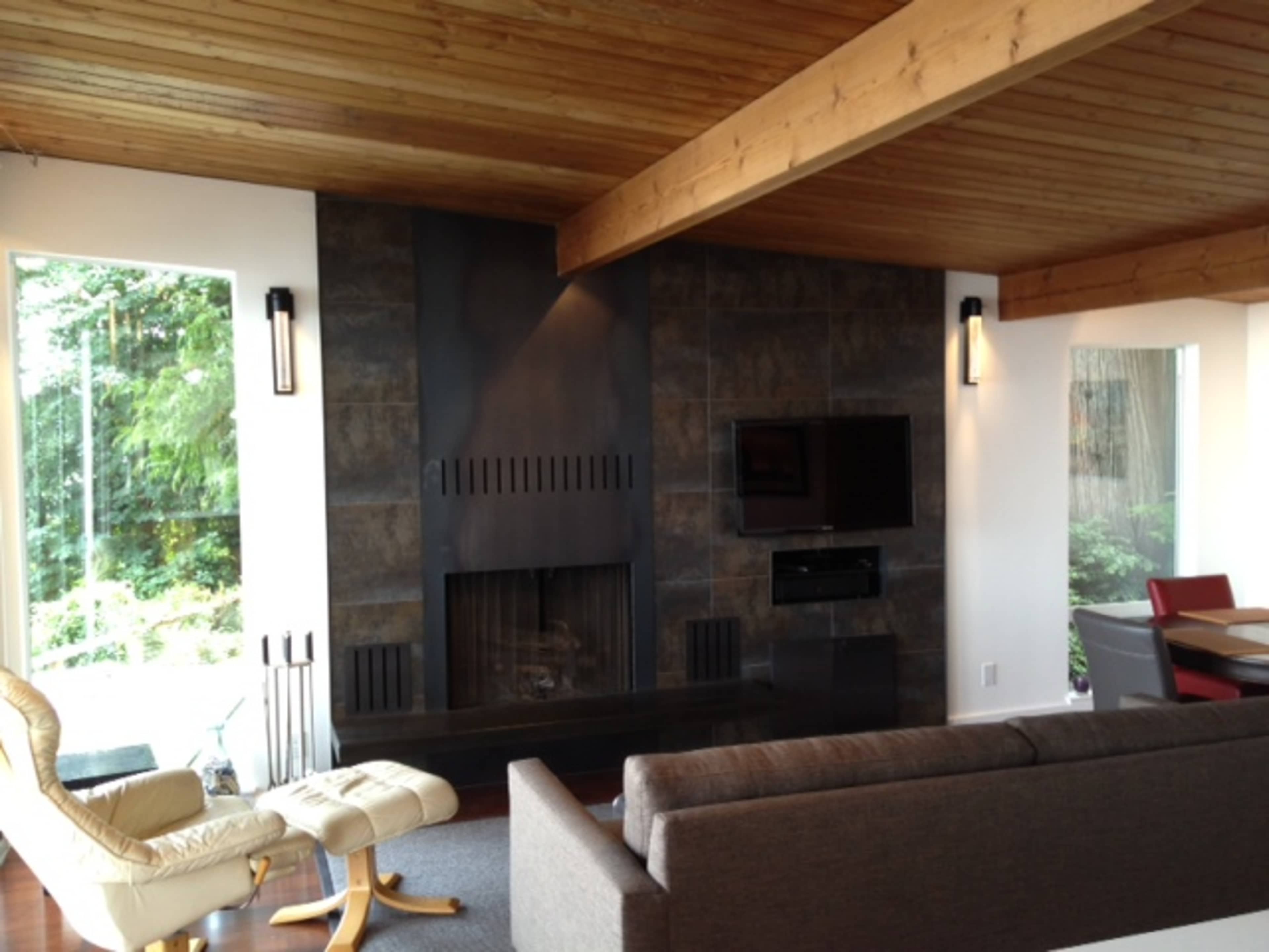 A modern living room featuring a stone fireplace, a flat-screen TV mounted on the wall, and large windows letting in natural light.