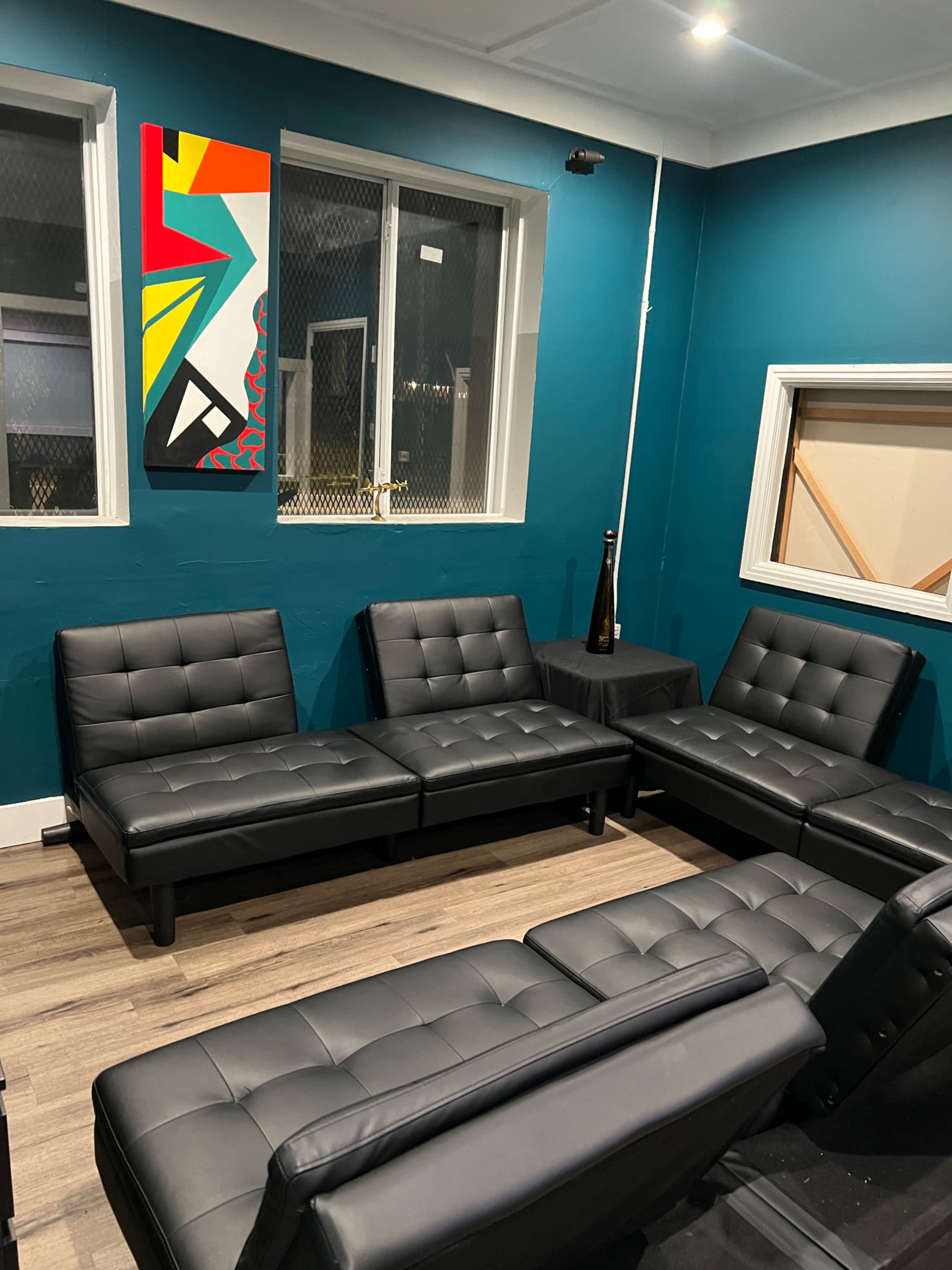 The image shows a small lounge area with black modular couches arranged around a table, against a teal wall featuring a colorful abstract painting.